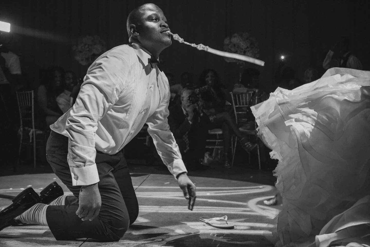 A man on his knees, wearing a white dress shirt and tie, is holding a lit cigar in his mouth with smoke trailing out, surrounded by an audience at a formal event.