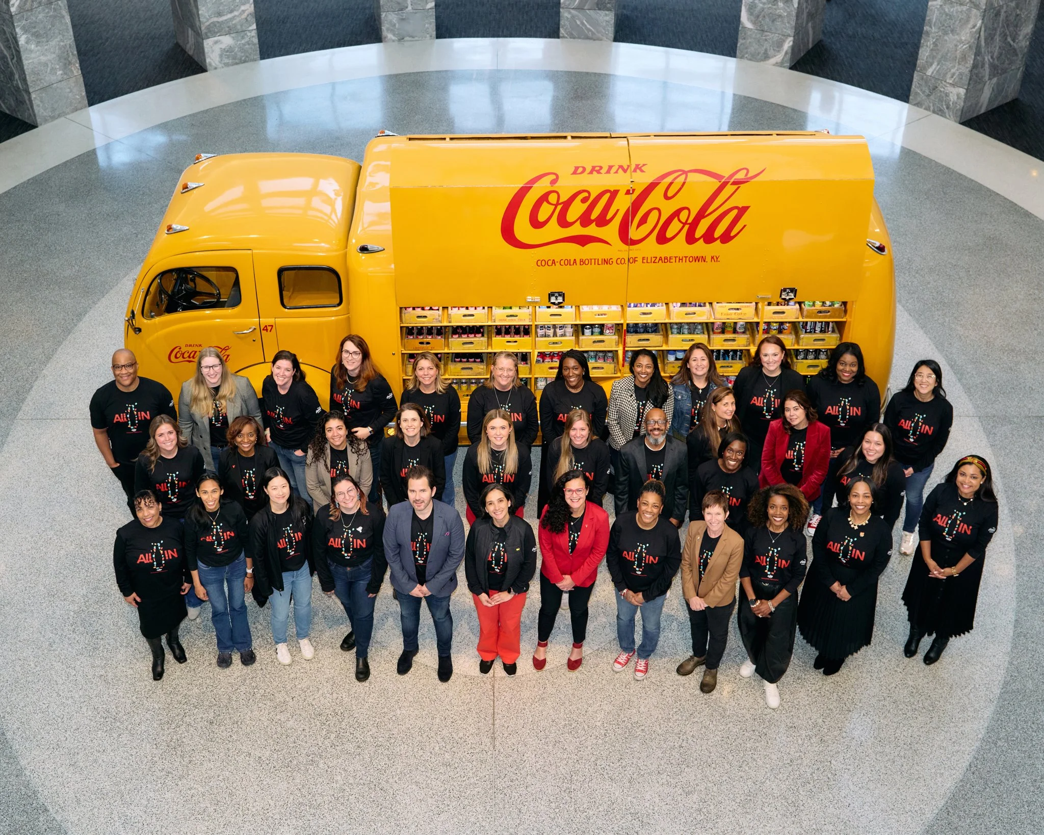A large group of diverse people standing in front of a yellow Coca-Cola delivery truck inside a building with a shiny floor and gray walls, smiling at the camera.