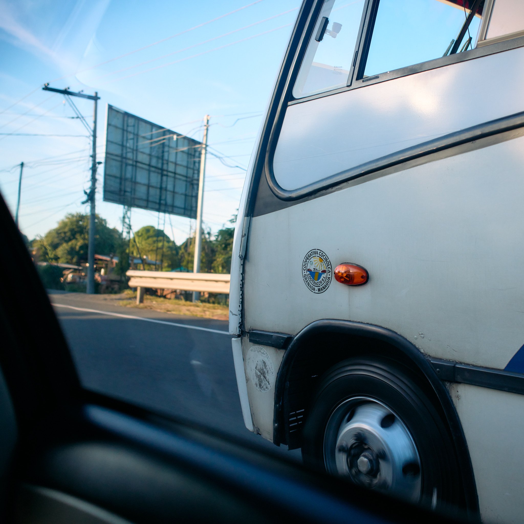 Close-up of a vintage white Volkswagen van with an emblem and orange side marker light, seen from a moving vehicle on the road during daytime.