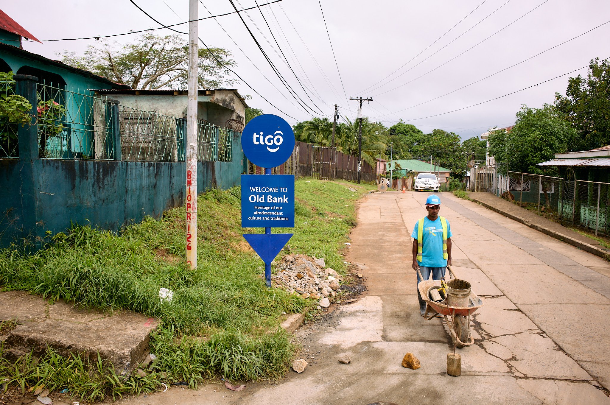 Street scene in Old Bank,  Blue fields  Nicaragua with a man pushing a wheelbarrow, blue sign welcoming visitors, green houses, trees, and a car at the end of the road.