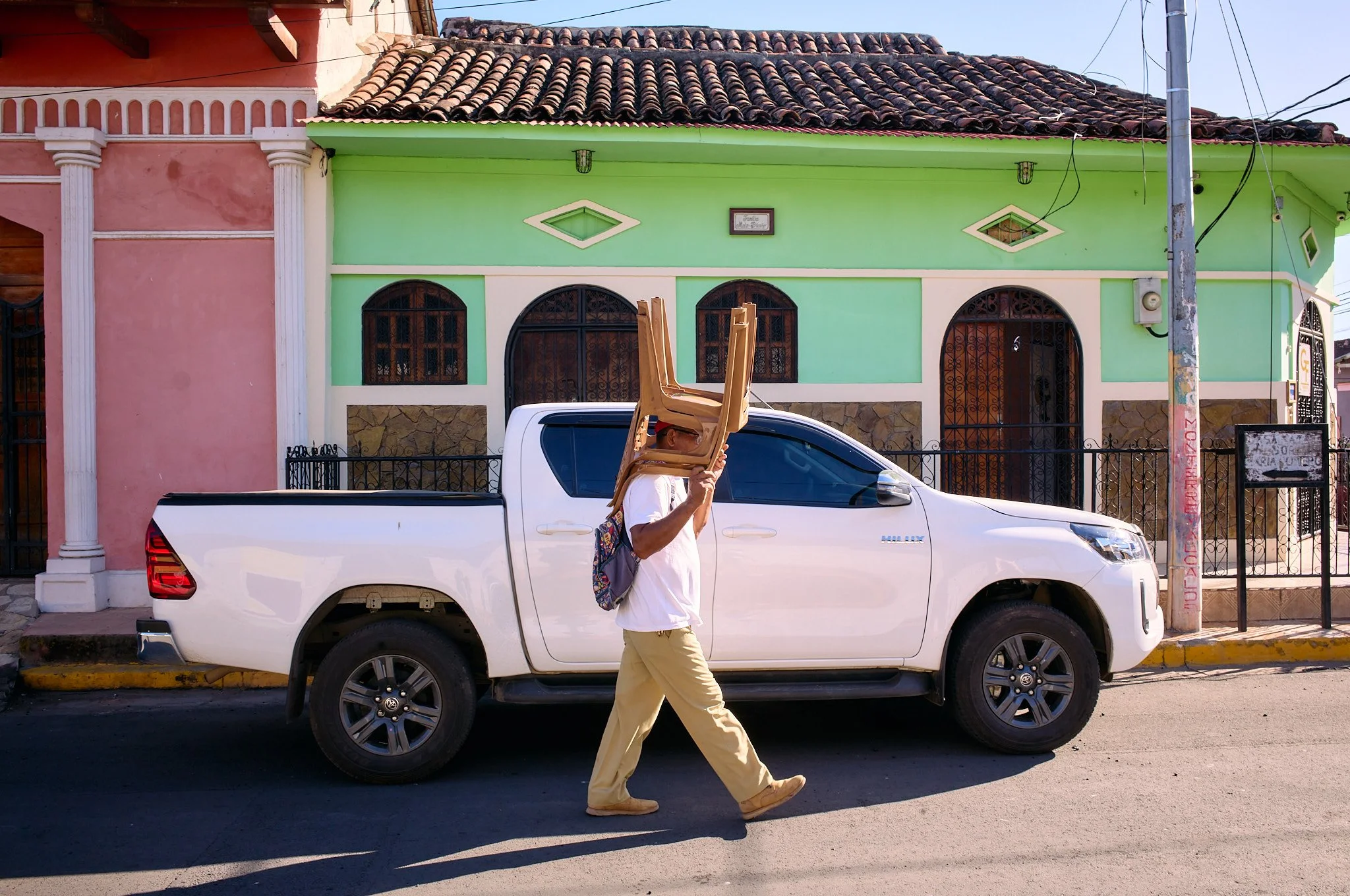 A man walking on the street carrying three stacked chairs on his shoulder in front of a white pickup truck and colorful houses.