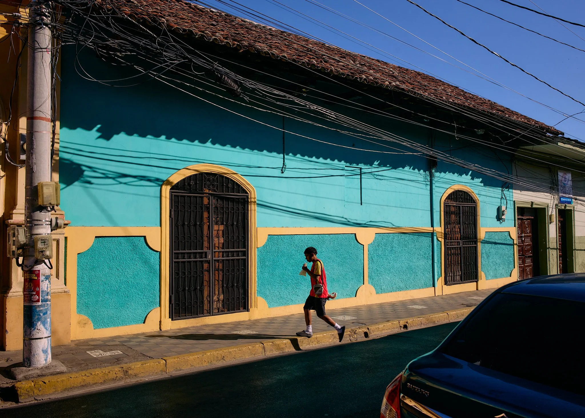 A young man in a red shirt and shorts walks along a sidewalk while looking at his phone in front of a colorful turquoise and yellow building under a bright blue sky, with parked car partially visible in the foreground.