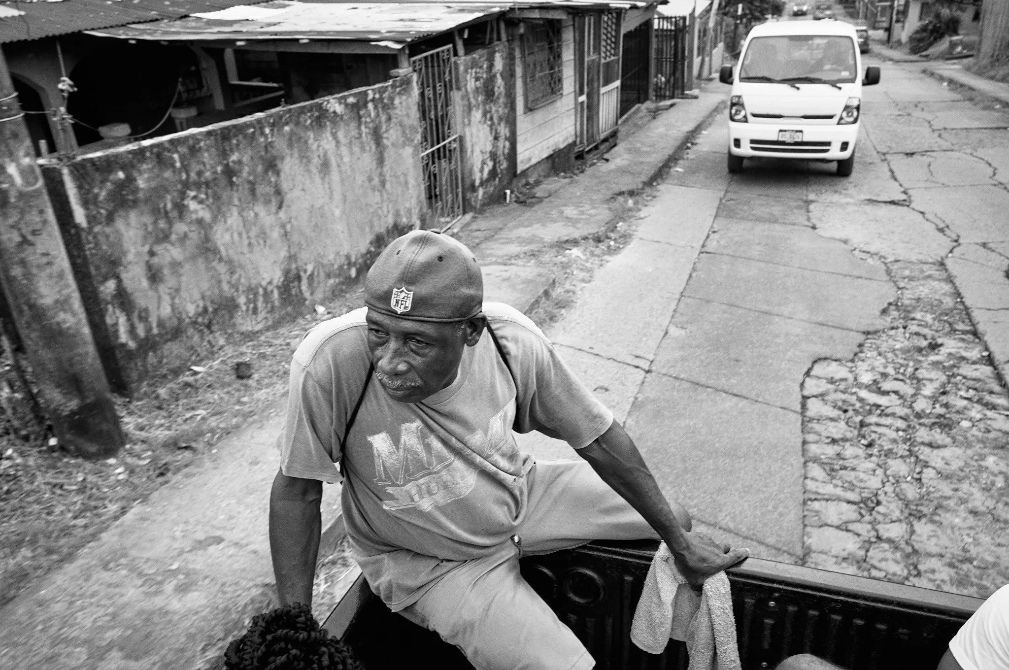 A man in casual clothing and a cap sitting on the back of a vehicle, with a street scene featuring a parked white van and a cracked sidewalk in the background.
