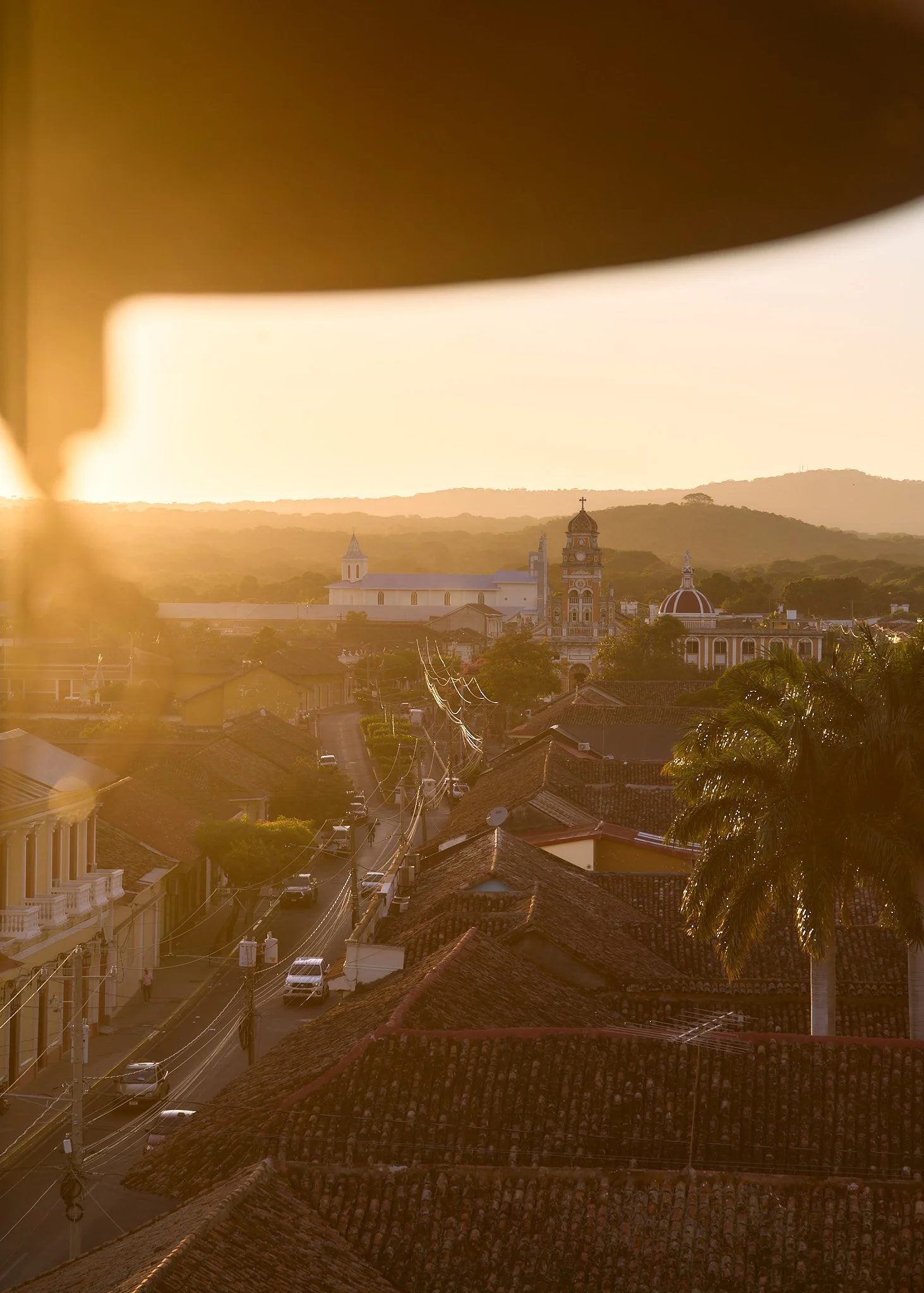 A cityscape at sunset of Granada Nicaragua with red tiled roofs, a palm tree, and church steeples in the background.