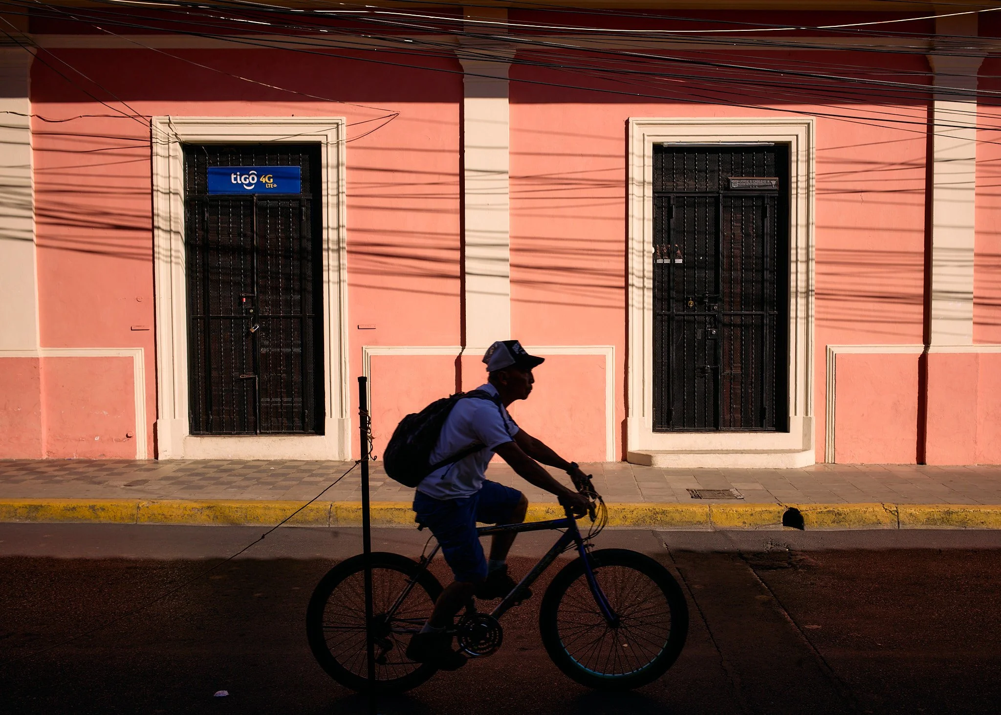 Silhouette of a person riding a bicycle in front of a pink building with barred windows, shadows of power lines cast on the wall.