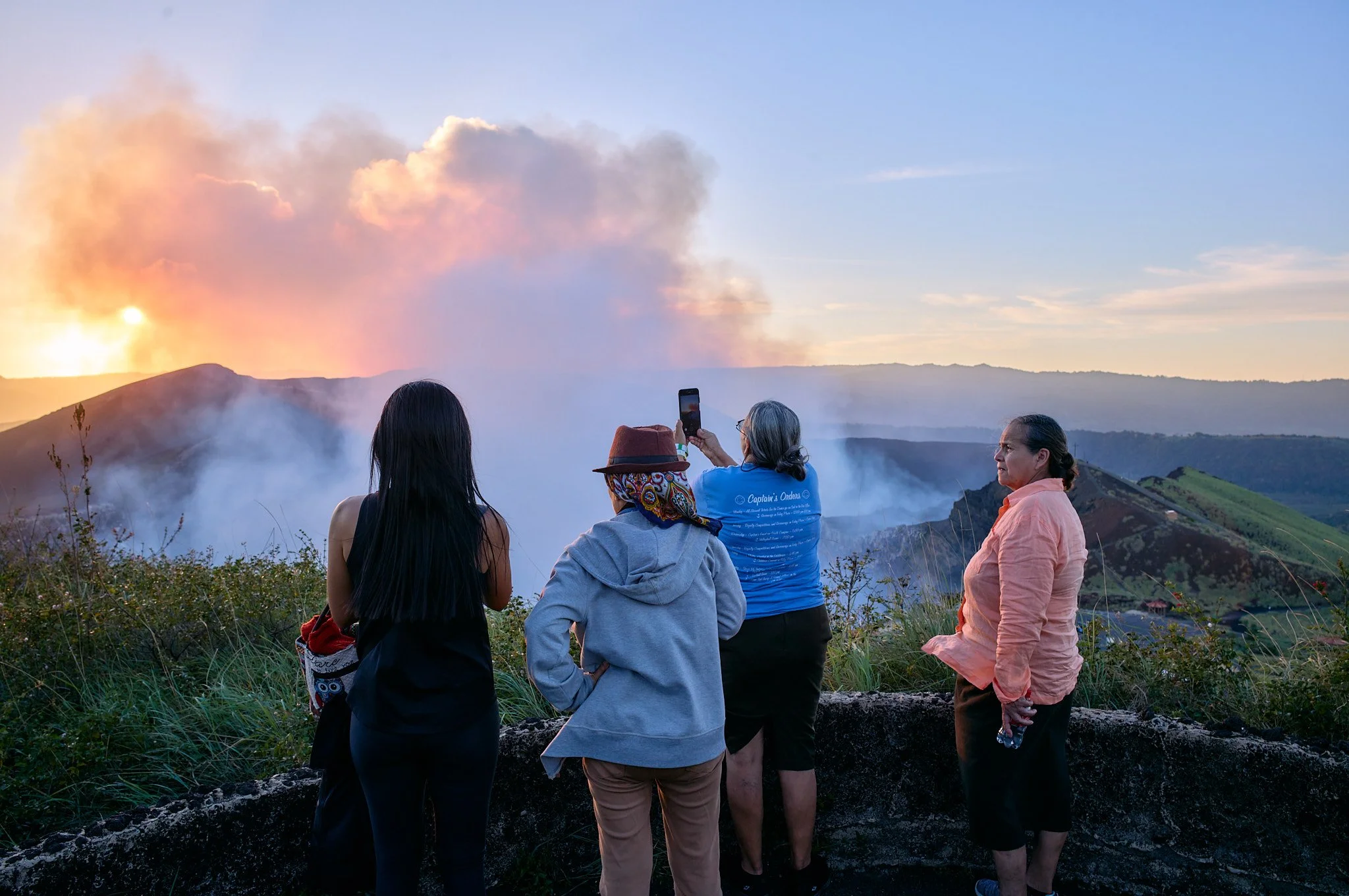 Four women observing a volcanic eruption at sunset, with one woman taking a photo with her phone. Smoke and ash billow from the volcano in the background.