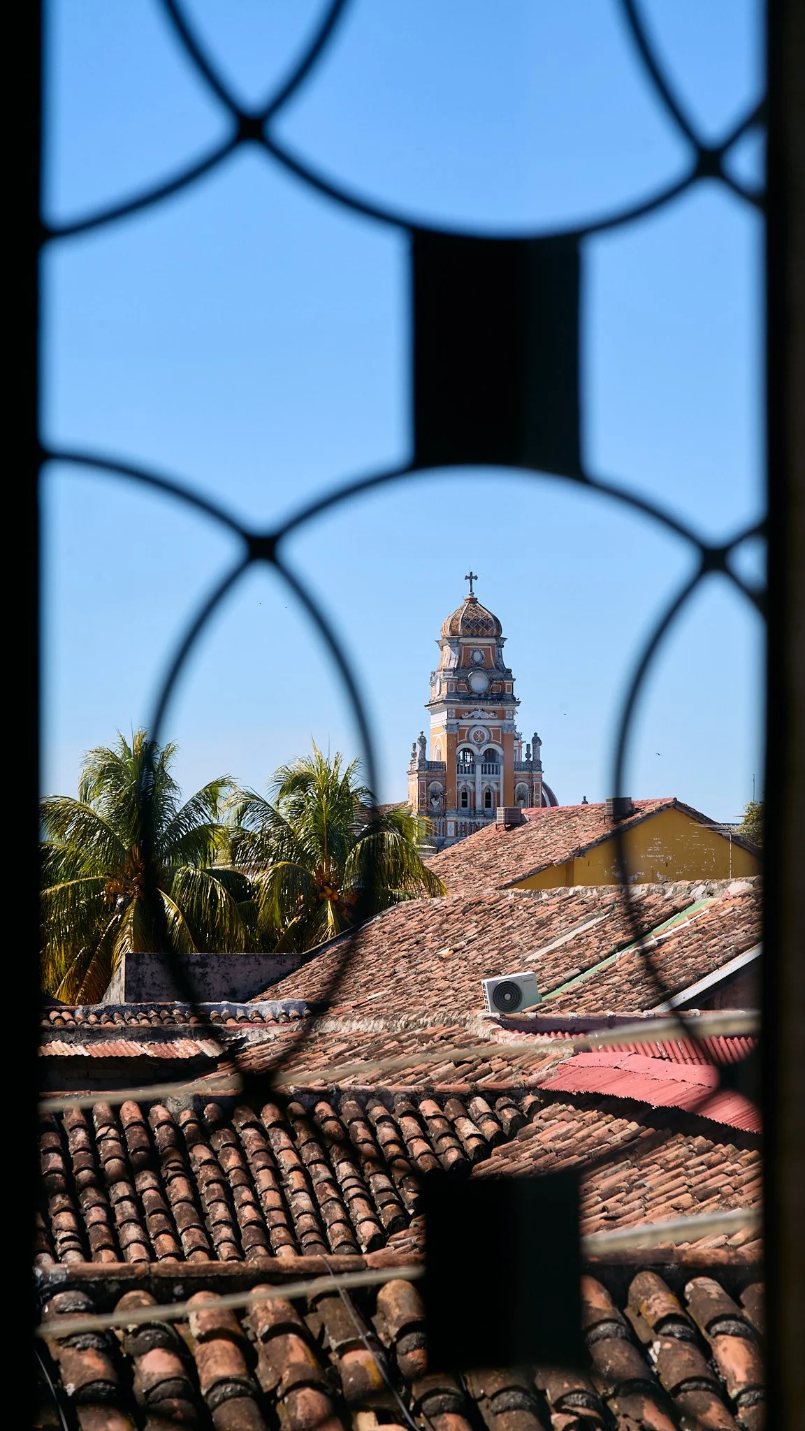 View of a church tower with a cross on top, seen through a decorative window grille, with palm trees and rooftops in the foreground.