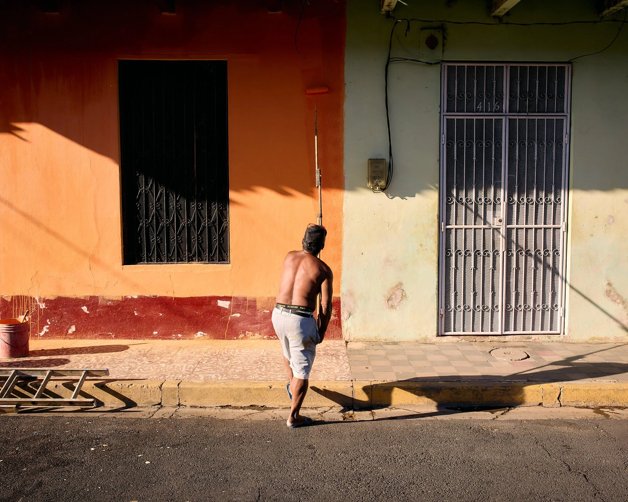 A shirtless man wearing gray shorts and blue shoes walking on a sidewalk in front of a building with a green and orange wall, two windows with metal bars, and a metal gate. The man is seen from the back, and the scene is lit by bright sunlight creati
