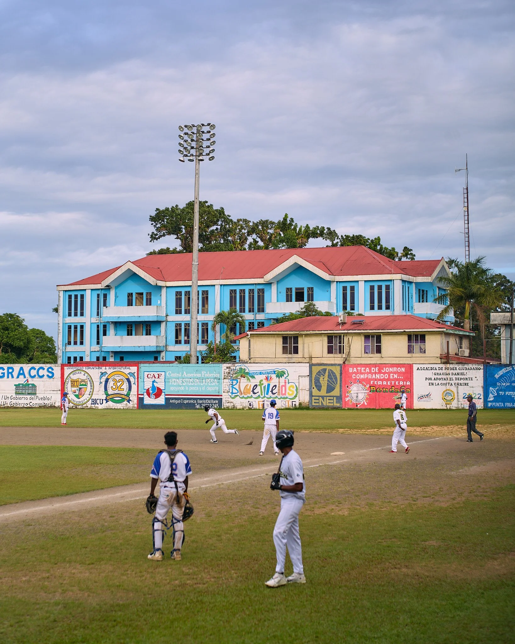 Baseball players on a field with a colorful building and advertisements in the background in blue fields Nicaragua 