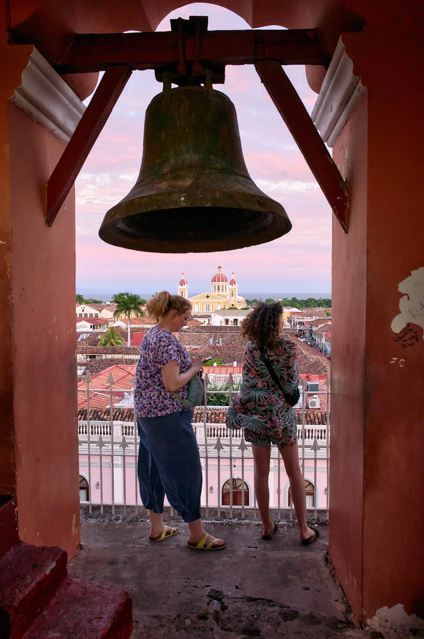 Two women standing in a bell tower overlooking the Granada, Nicaragua cityscape with a colorful church in the background, during sunset.