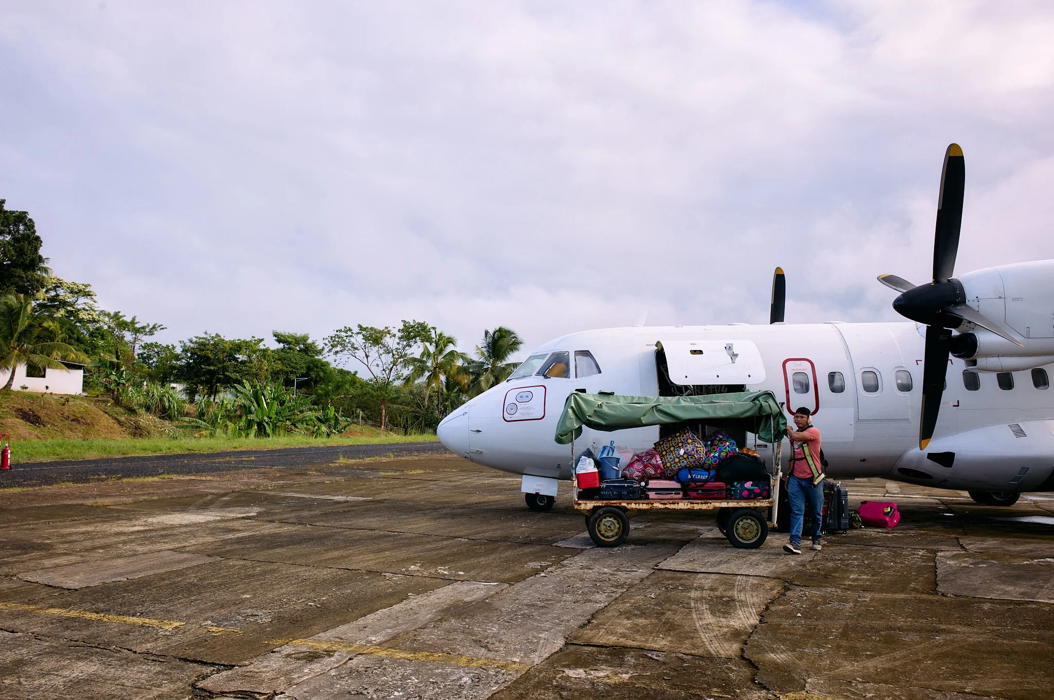 Person loading luggage onto a small aircraft at Bluefeilds Nicaragua airport tarmac with lush green trees and cloudy sky in the background.