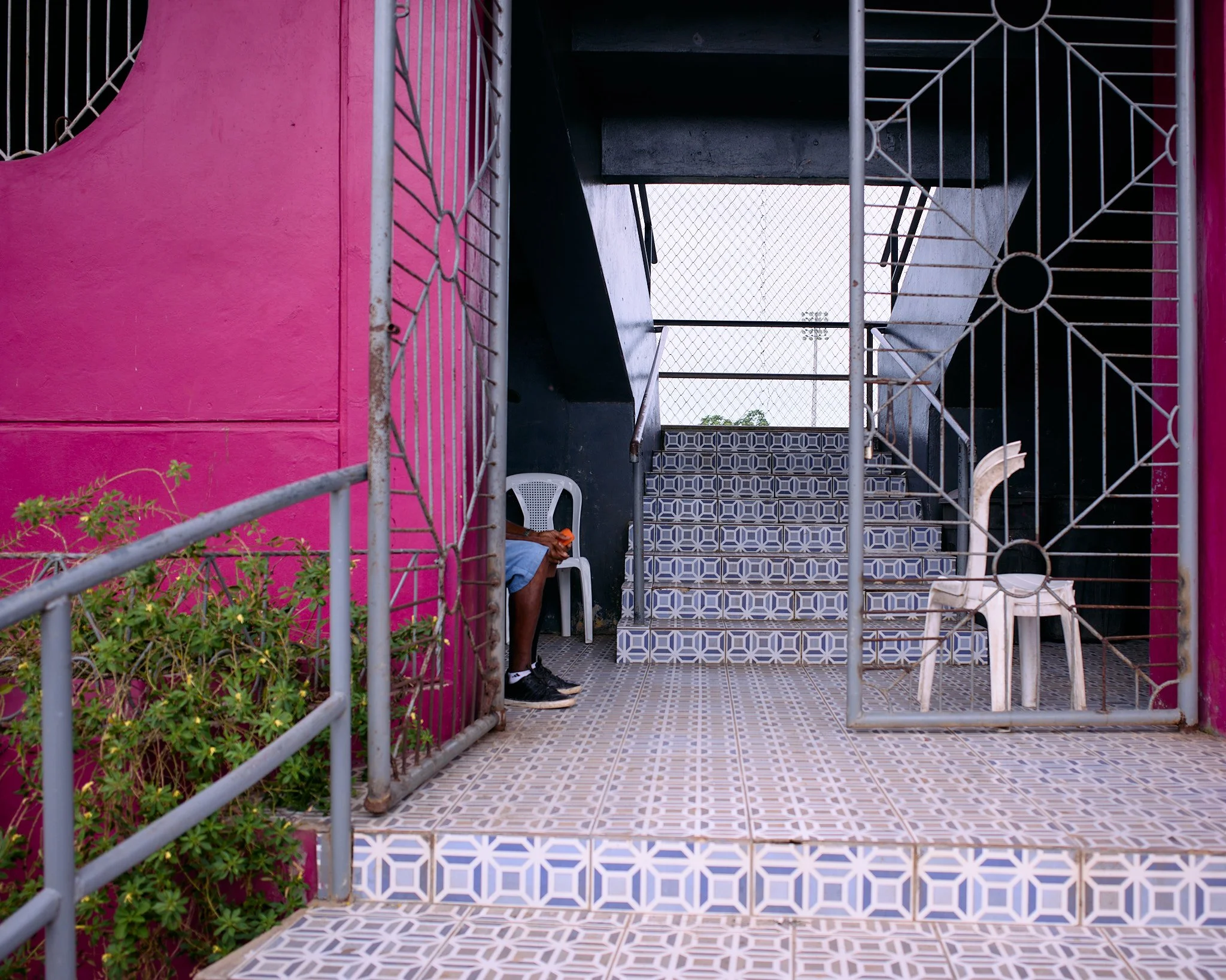 A small outdoor area with a pink wall, tiled flooring, and a staircase. There is a white plastic chair with a person sitting on it, partially hidden, and another empty white plastic chair that looks like a toy giraffe. A metal gate surrounds the spac