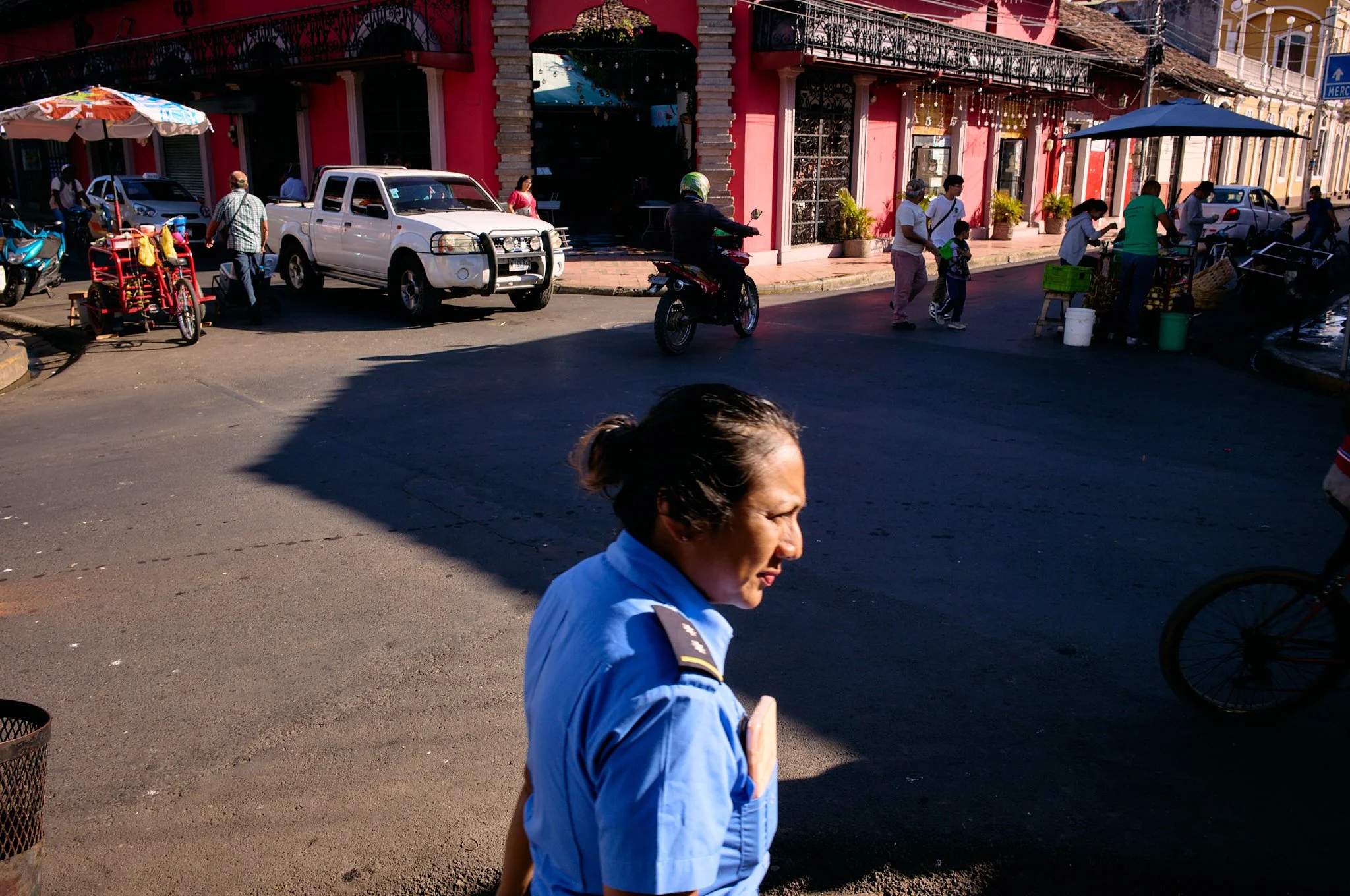 A woman in a blue uniform standing in the foreground on a street with buildings, cars, motorcycles, people, and vendors in the background.