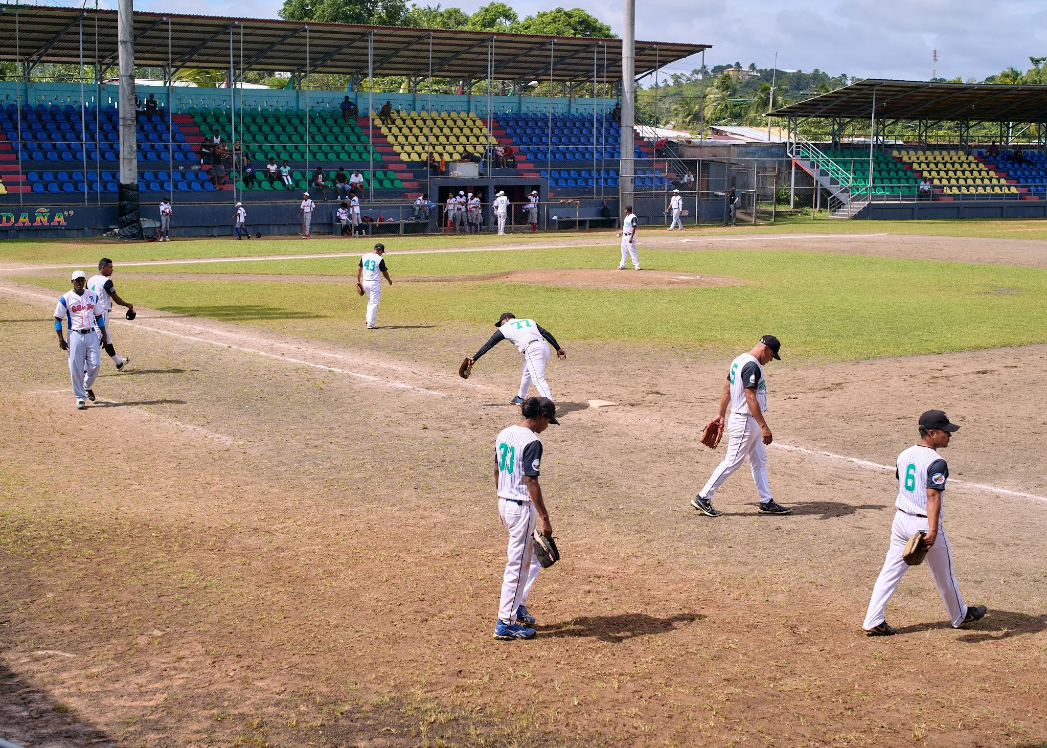 A baseball game with players walking on the field and others in the dugout, surrounded by colorful stands and a partially cloudy sky.