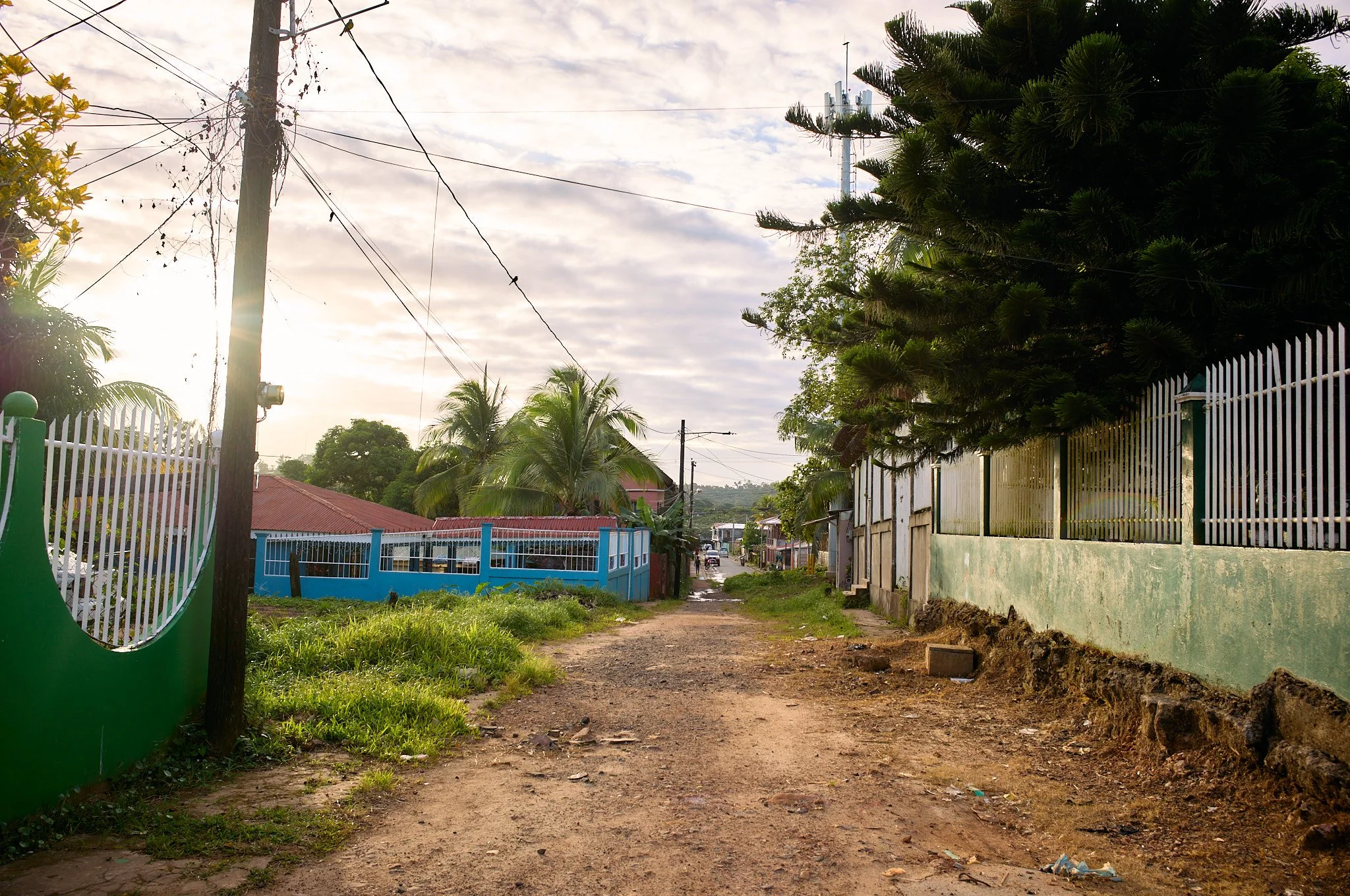 A dirt road in a residential neighborhood with houses and fencing on both sides, palm trees, and utility poles with overhead wires under a partly cloudy sky.