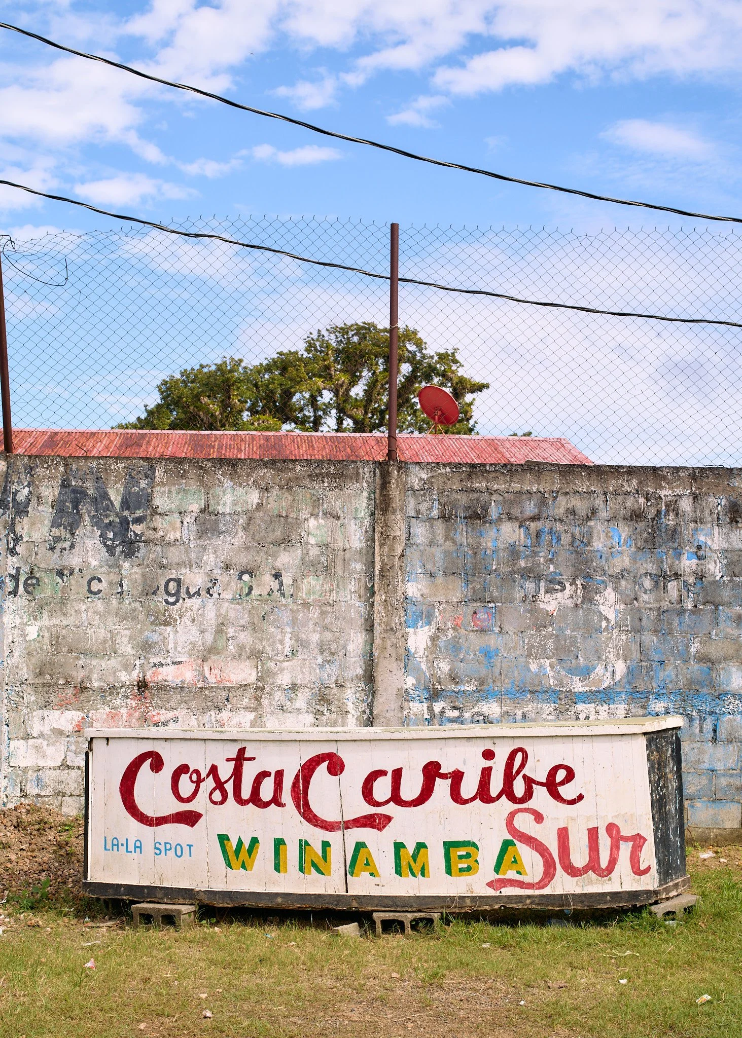 A weathered wooden sign in front of a concrete wall with faded graffiti, with a blue sky and clouds above in blue fields Nicaragua 