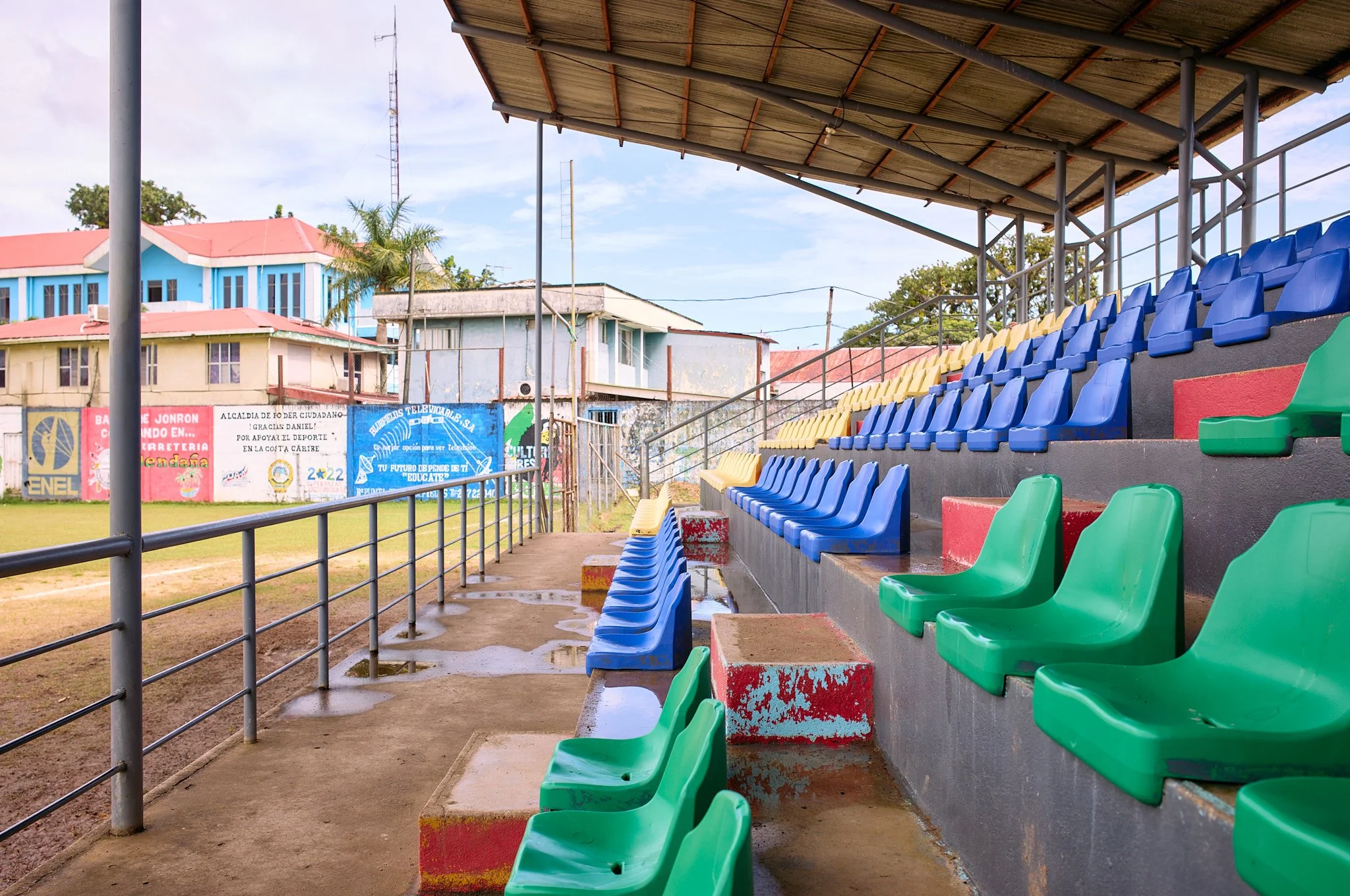 Empty bleachers with green, yellow, and blue seats under a roof at a soccer field, with murals and buildings in the background in blue fields Nicaragua 