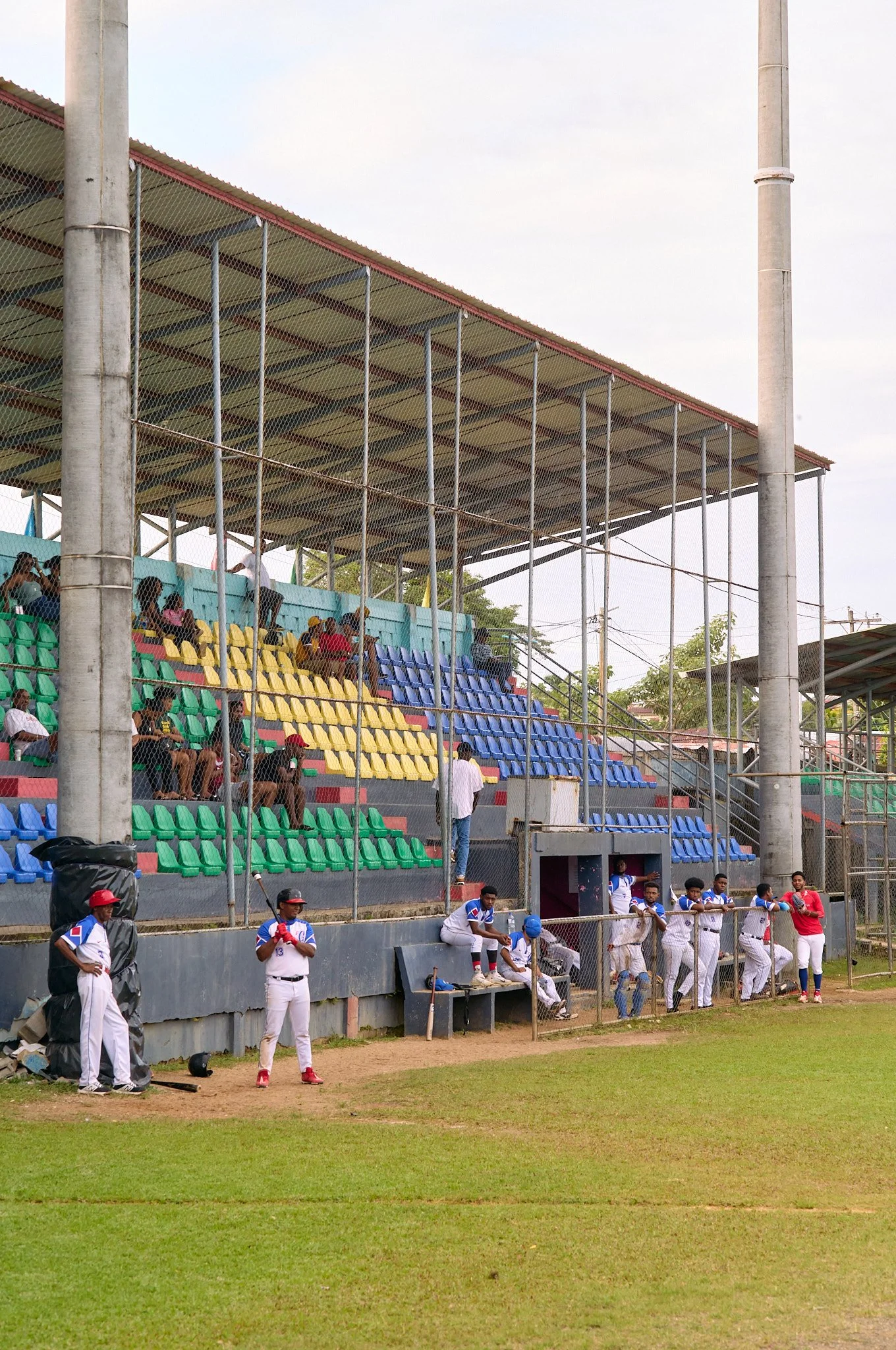 Baseball field with players and spectators, some players are sitting on the bench while others are standing near the dugout. Spectators are seated in colorful bleachers under a metal roof, and some are watching from behind a chain-link fence.