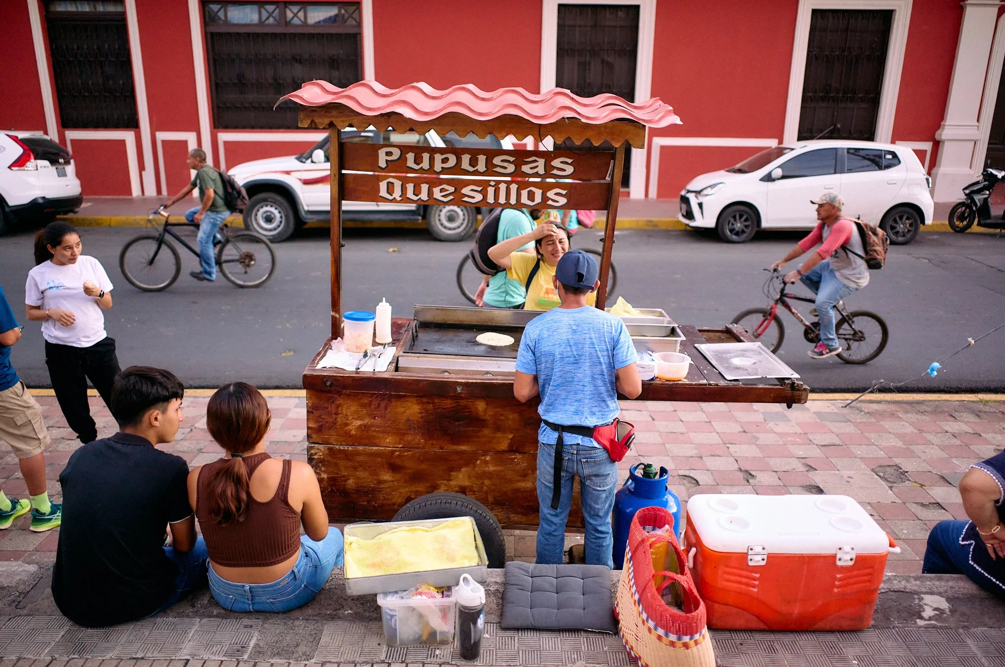 Street scene featuring a taco stand named 'Pupusas Quesillos' with a woman working behind the counter, a man in a blue shirt placing an order, and several people sitting and walking nearby. Cars and bicycles are visible on the street, with a red buil