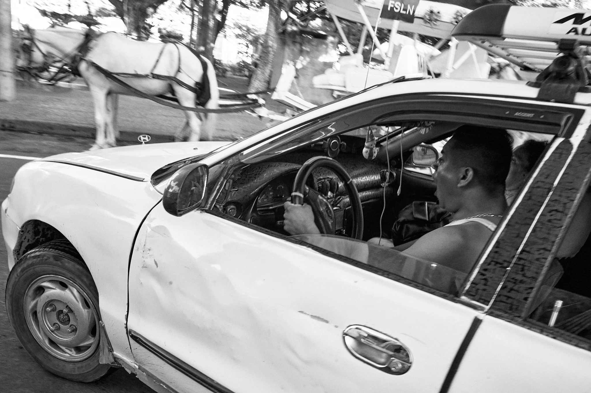 A black and white photo of a man driving a damaged car with a police badge on the hood. There is a horse in the background and some boats on a trailer.