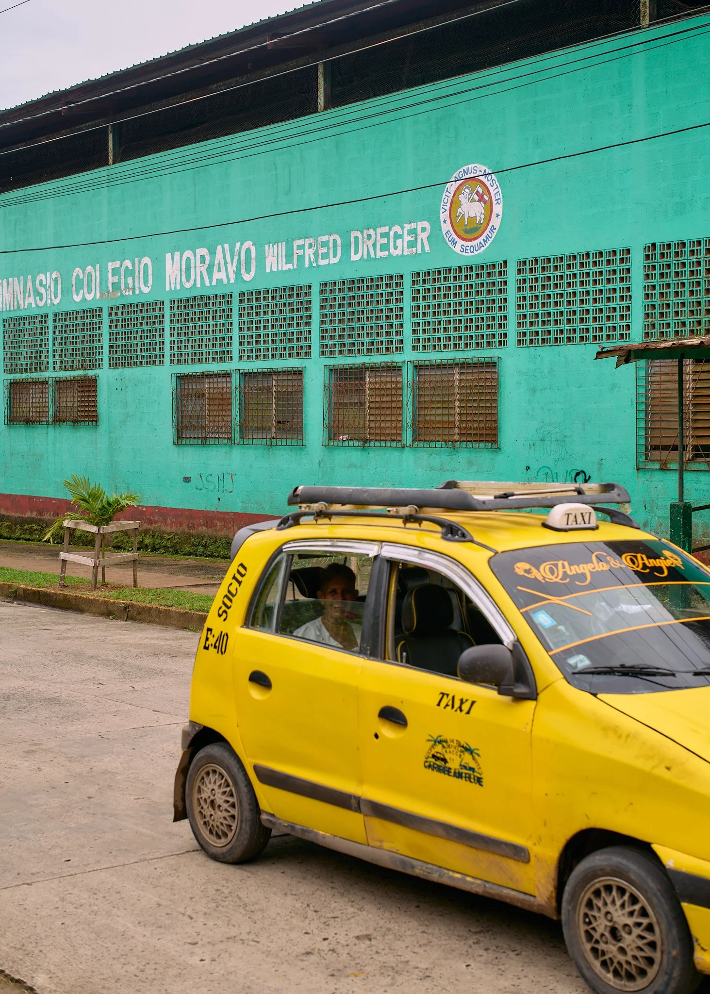 A yellow taxi parked on the street in front of a teal school building with the sign 'Gimnasio Colegio Moravo Wilfred Dreger' and a school emblem.