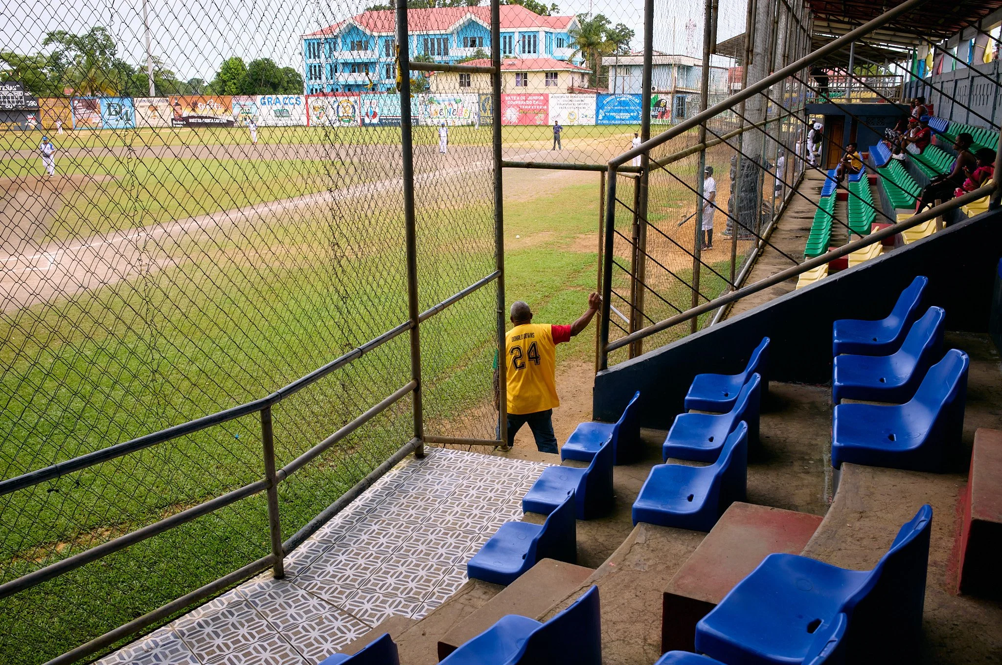 A baseball game at a local field in blue fields with spectators in the stands, one man wearing a yellow jersey with the number 24 is standing at the gate.