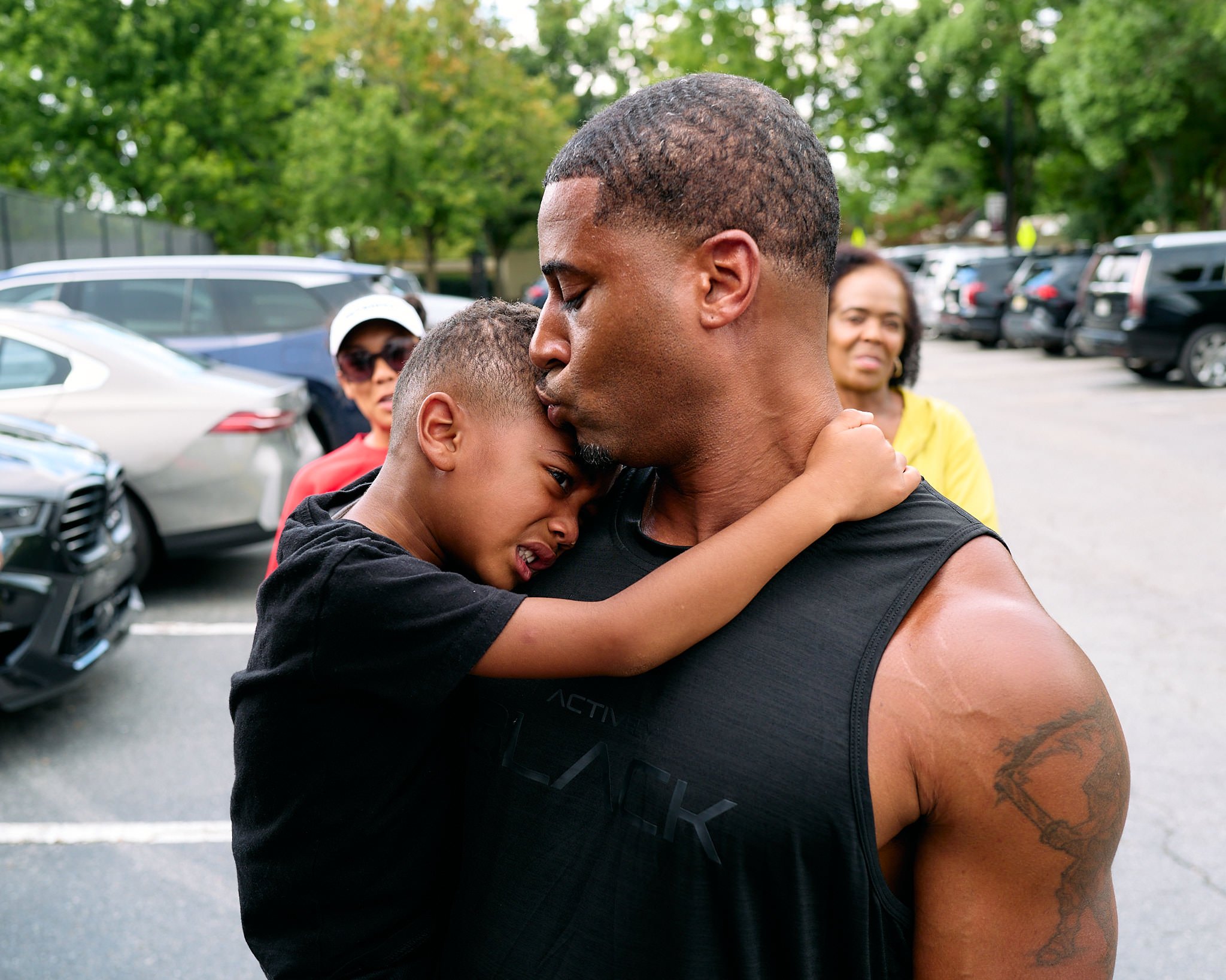 An emotional moment showing a young boy hugging and kissing a man, possibly his father, in a parking lot with trees and several parked cars in the background. Two women are observing in the background.