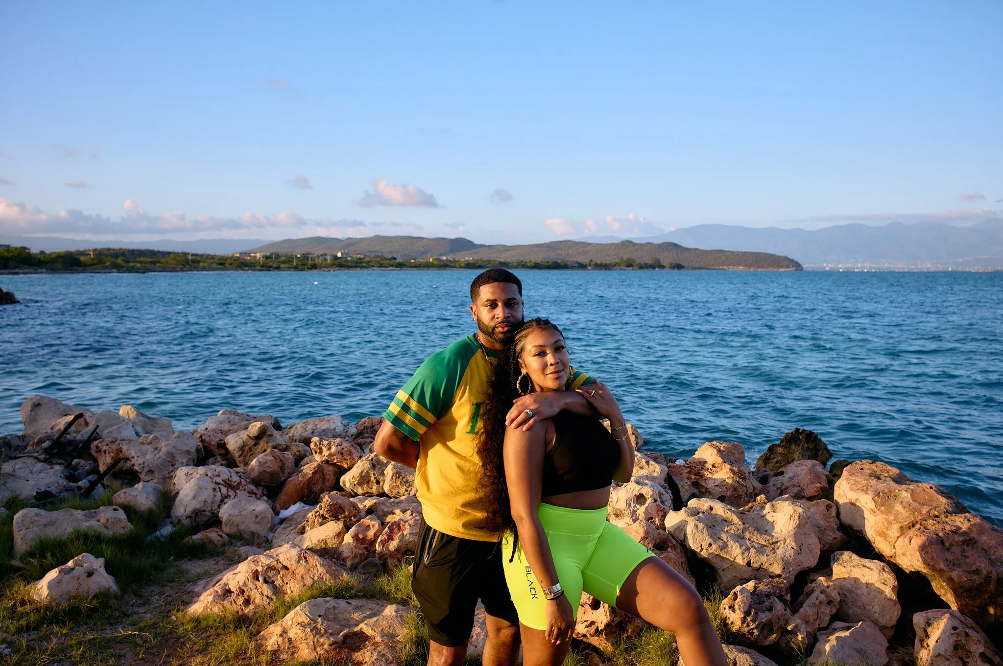 Devale and Khadeen Ellis posing by the rocky shoreline near a body of water with mountains in the background. The man is wearing a green and yellow sports shirt and black shorts, and the woman is dressed in a black top and bright yellow shorts.