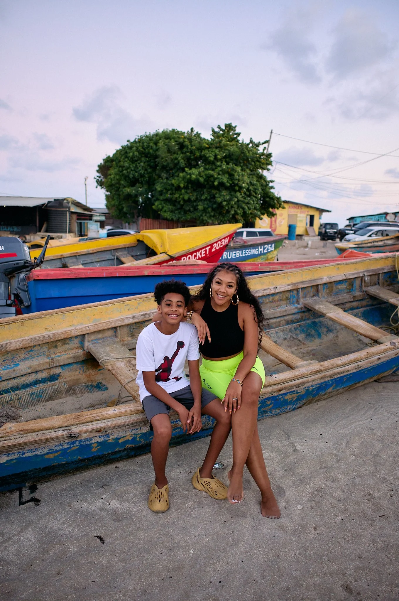 A woman and a boy sitting on a wooden boat on the beach, smiling at the camera, with colorful boats and a tree in the background.