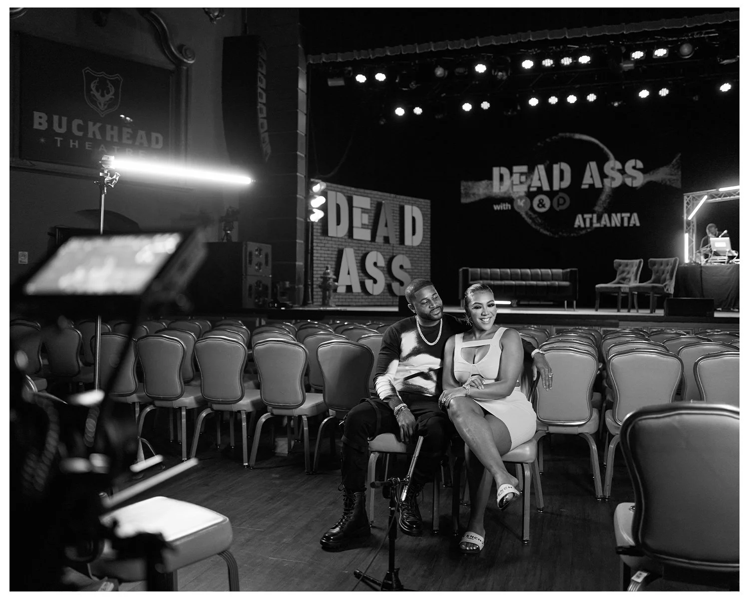 A black and white photo of a theater stage with the Ellises sitting in the audience. The stage backdrop has the text 'DEAD ASS' with 'with' and 'ATLANTA' below. Empty chairs are on the stage, and the seating area is mostly empty with rows of chairs.