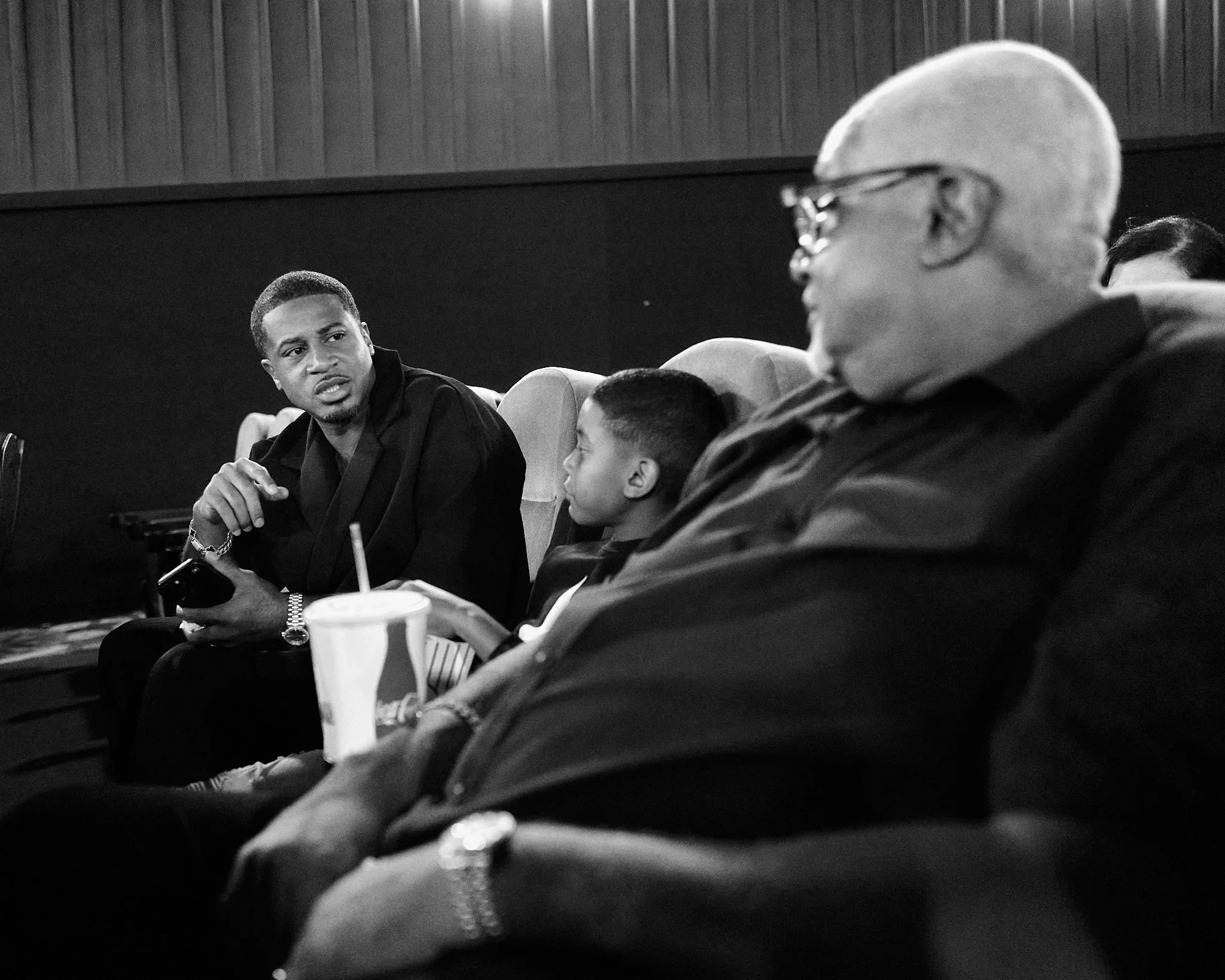 A black-and-white photo of three people seated in a theater or cinema. A young man on the left appears to be talking or explaining something. An older man on the right is looking down at his phone, and a young boy in the middle is looking at the youn