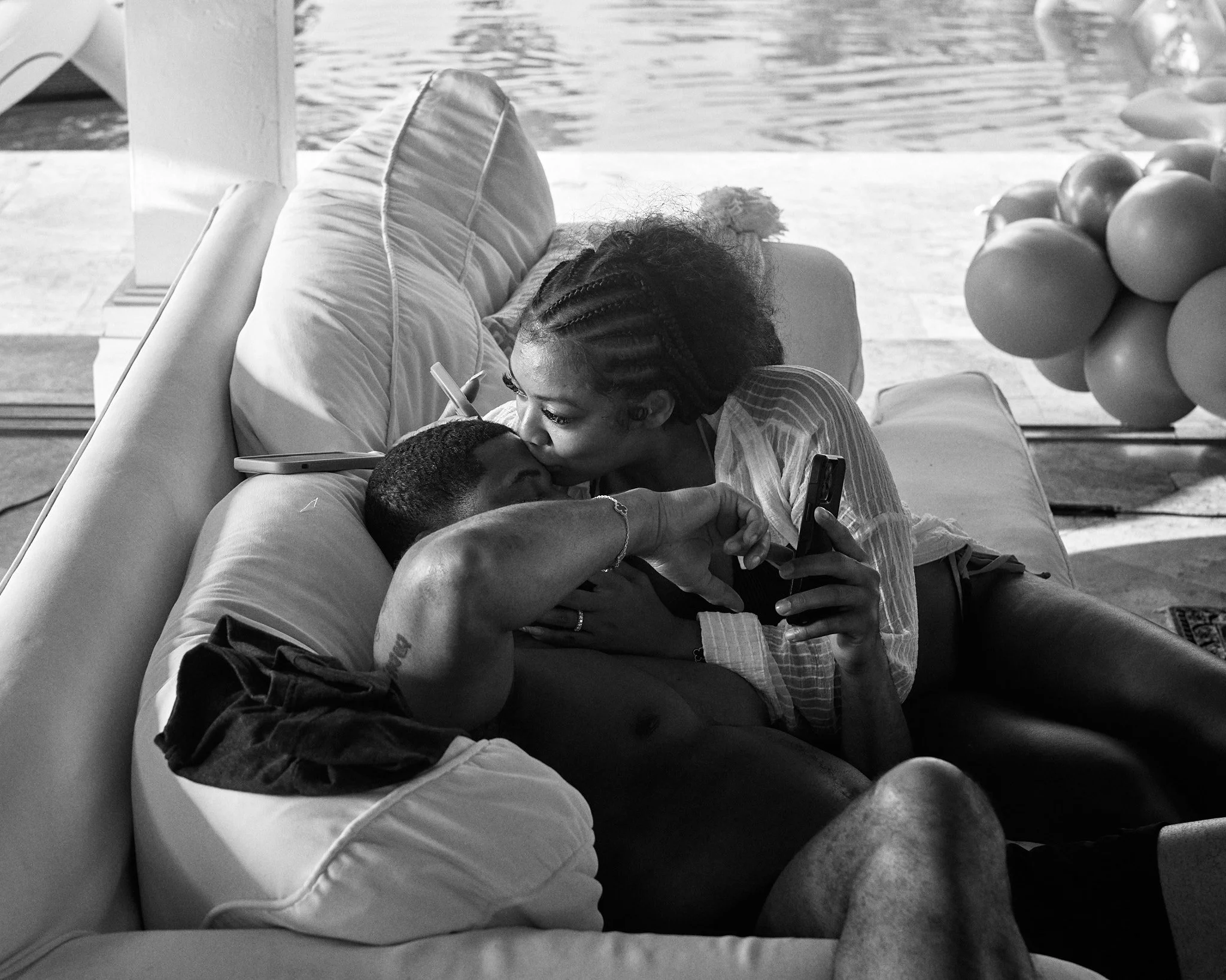 A woman and a man are sharing an intimate moment on a couch. The woman is leaning over and kissing the man on the forehead while holding a phone.
