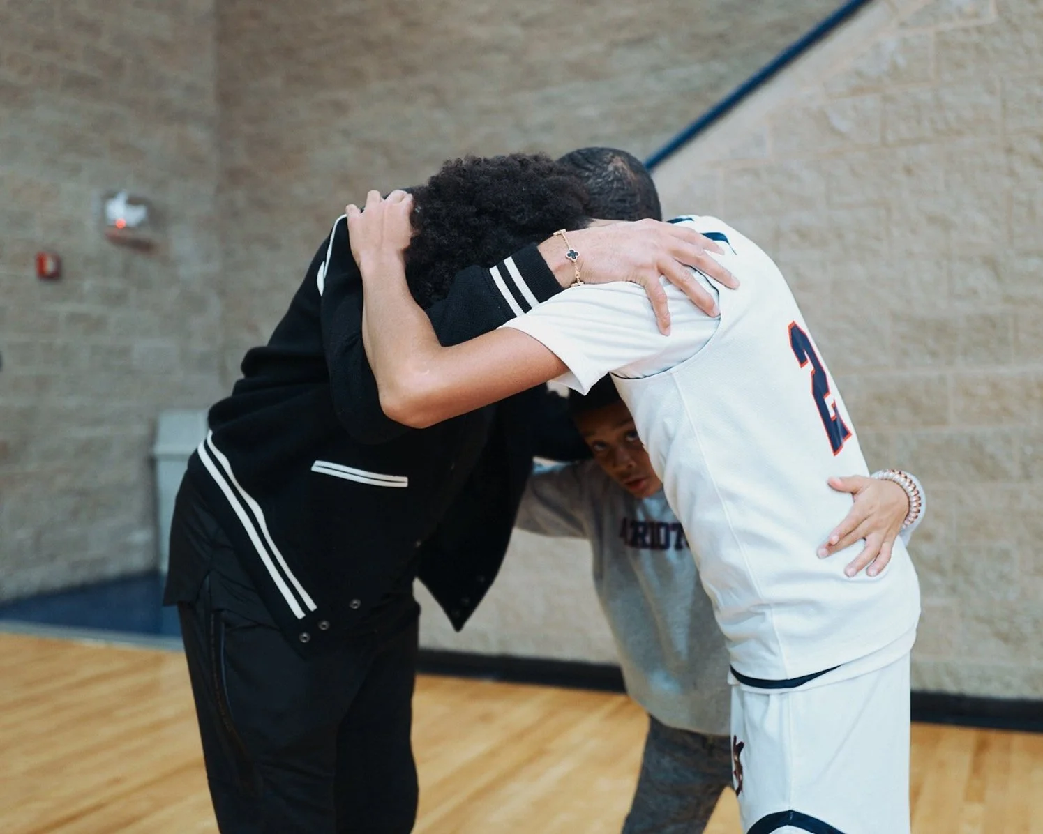 Two people hugging each other tightly, one in a black hoodie and one in a white sports uniform with the number 2, in a gymnasium.
