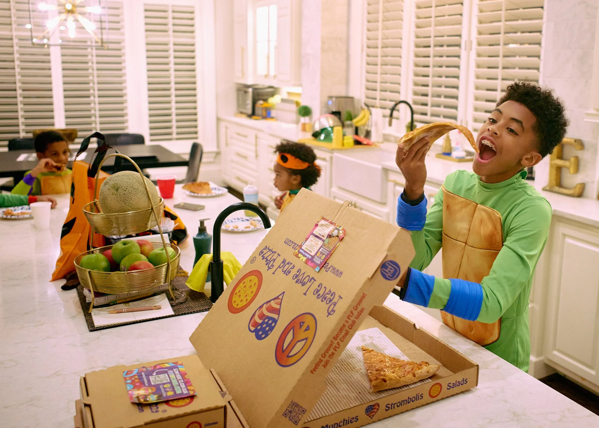 Children dressed as superheroes having a pizza party in a kitchen. One child is about to eat a slice of pizza, and the other children are sitting at a table with food and drinks.