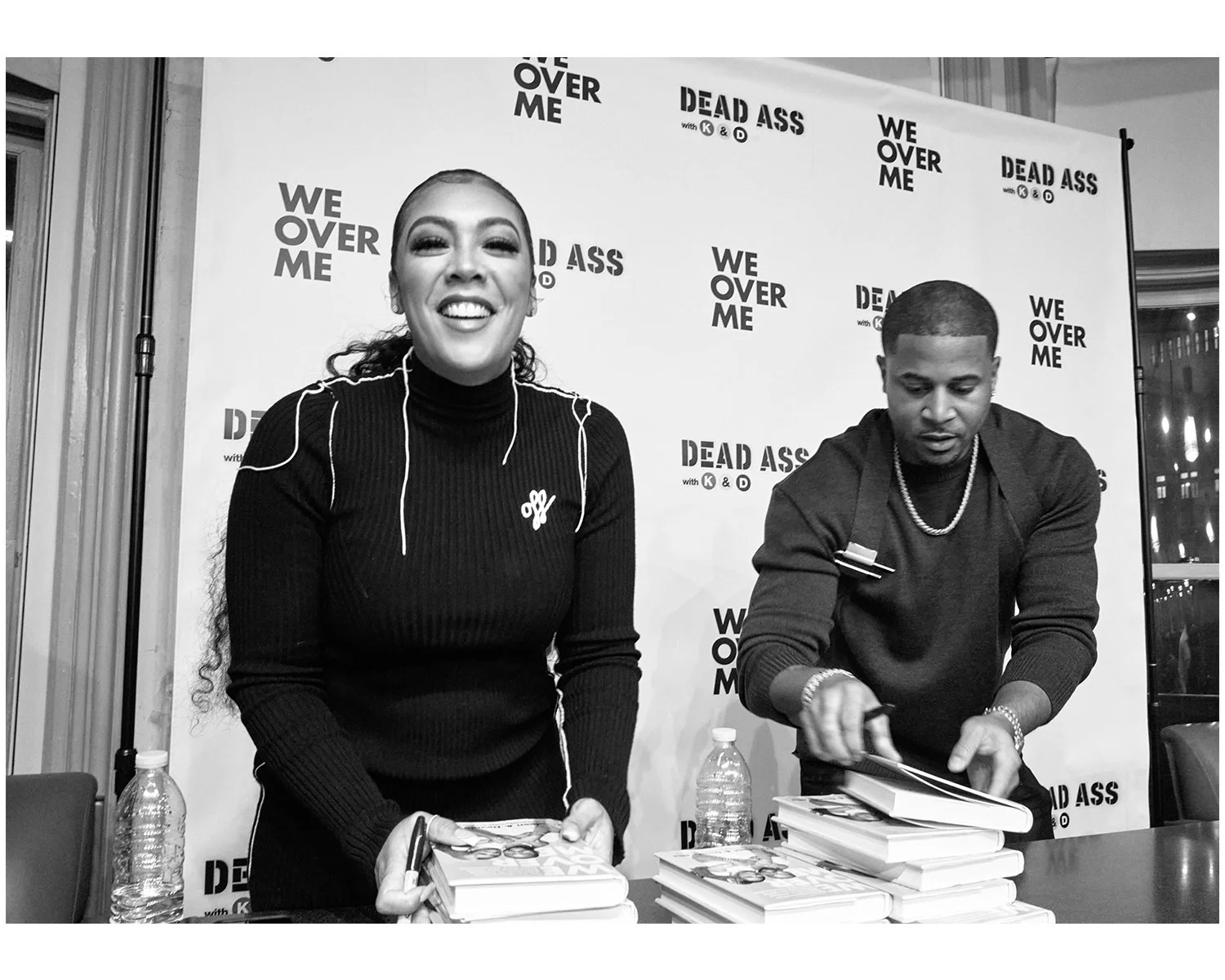 The Ellises both signing books at a promotional event with a backdrop reading 'DEAD ASS with K & D' and 'WE OVER ME.'