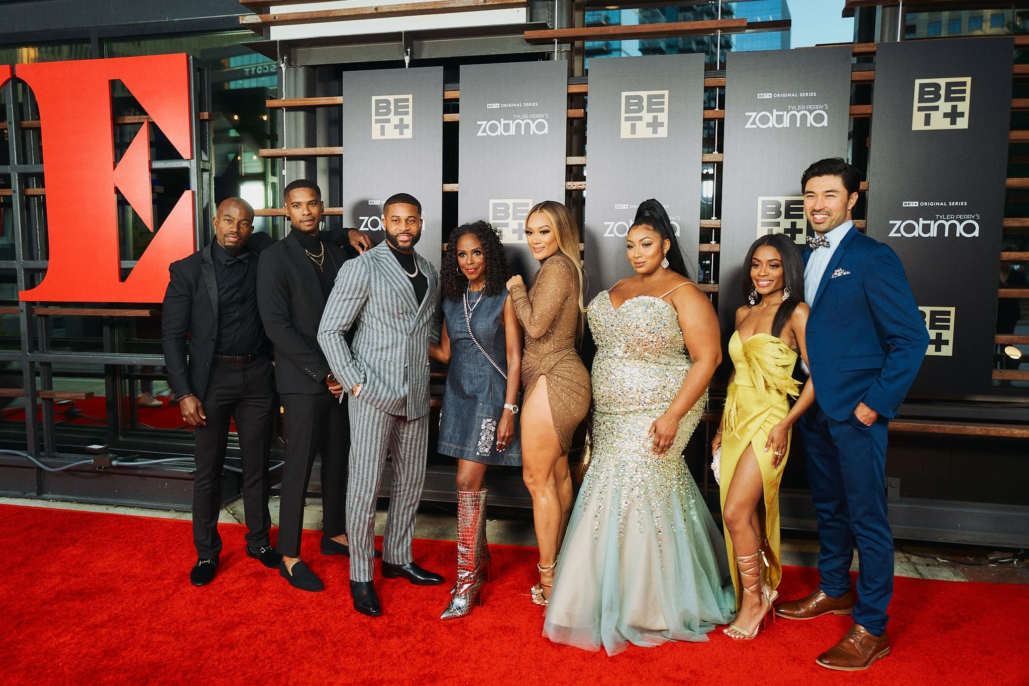 Group of nine people in formal attire posing on a red carpet in front of promotional banners for the TV series 'Zatima' and 'BET+'.