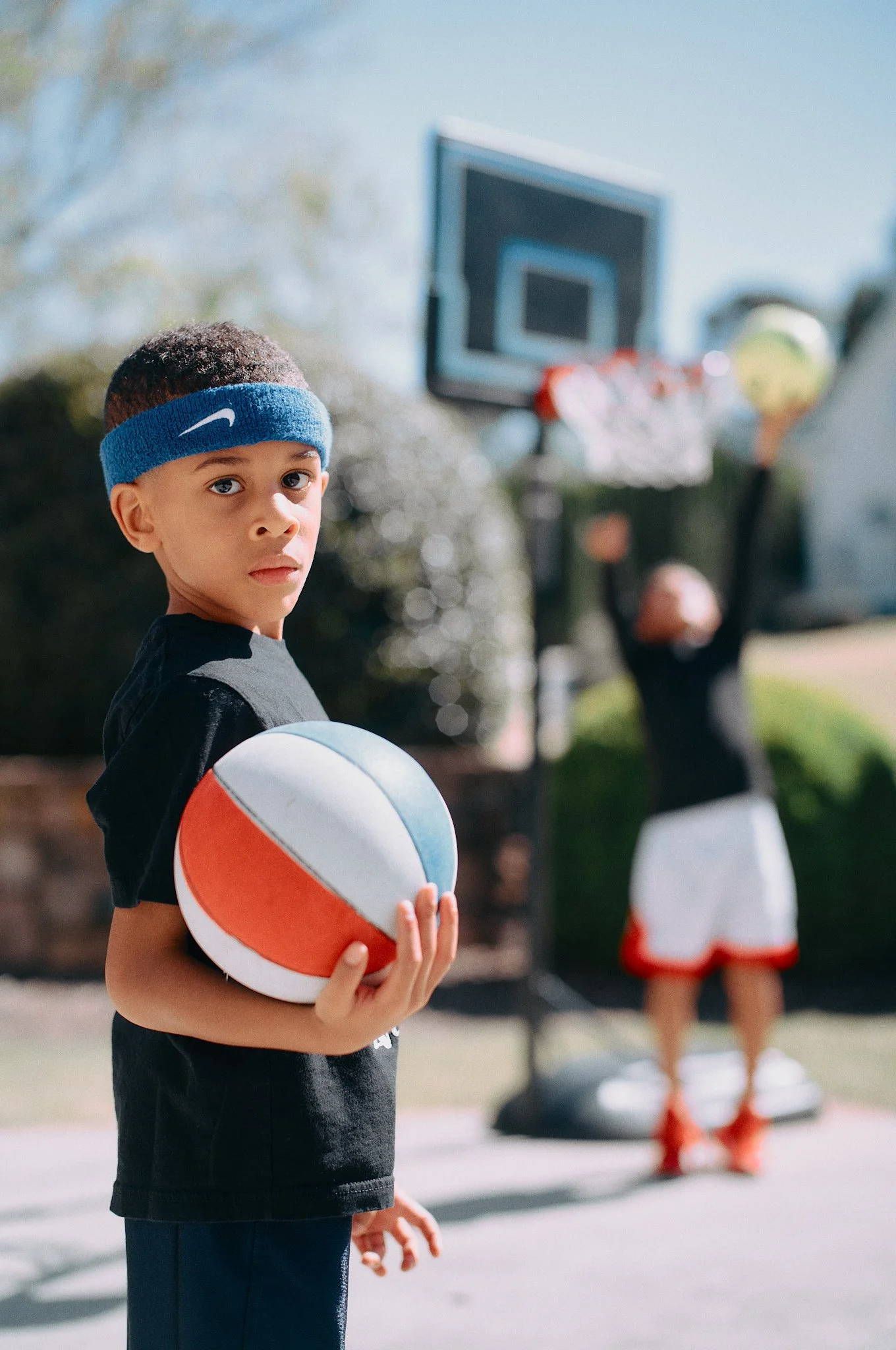 Young boy wearing a blue Nike headband holding a basketball on an outdoor basketball court, with another person playing basketball in the background.