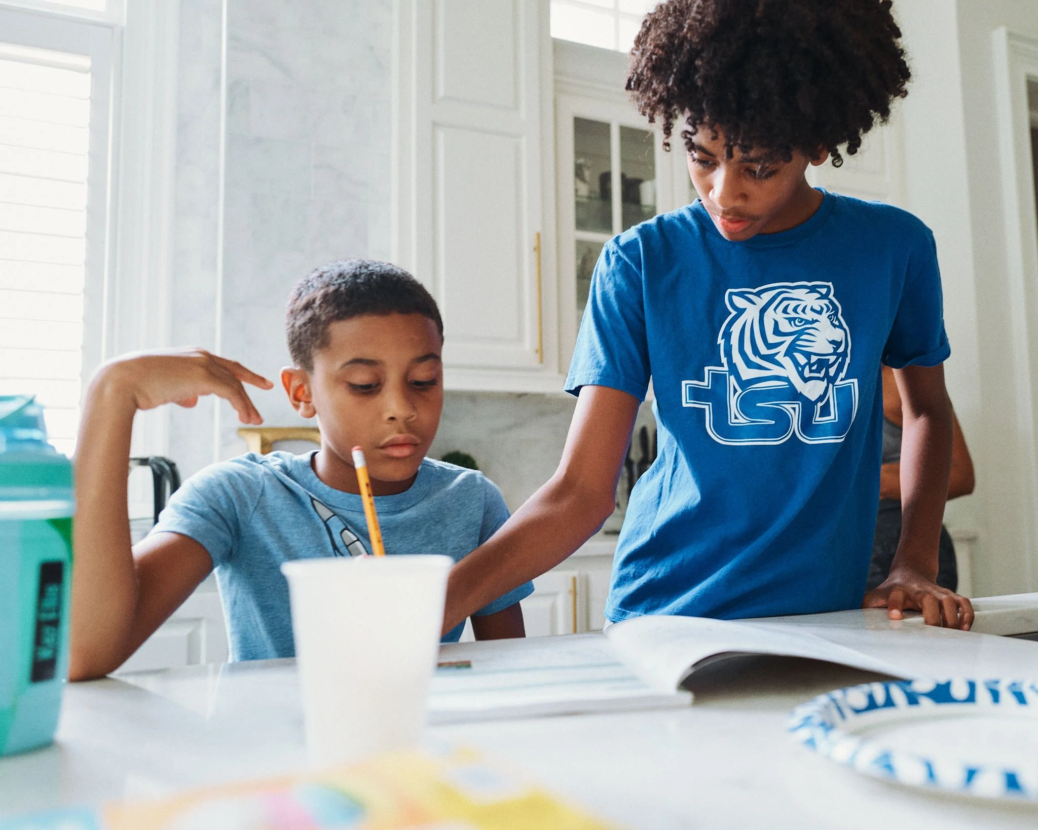 Two children, a boy and a girl, are studying at a kitchen table. The girl is pointing at a book while the boy looks focused. The girl is wearing a blue LSU t-shirt and the boy is wearing a light blue shirt. There are school supplies on the table.