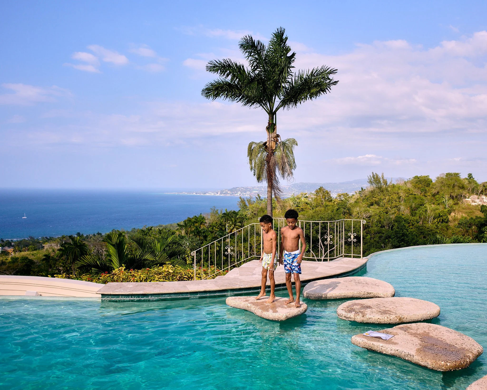 Two boys in swim trunks standing on stepping stones in a swimming pool with ocean view and palm trees in the background.