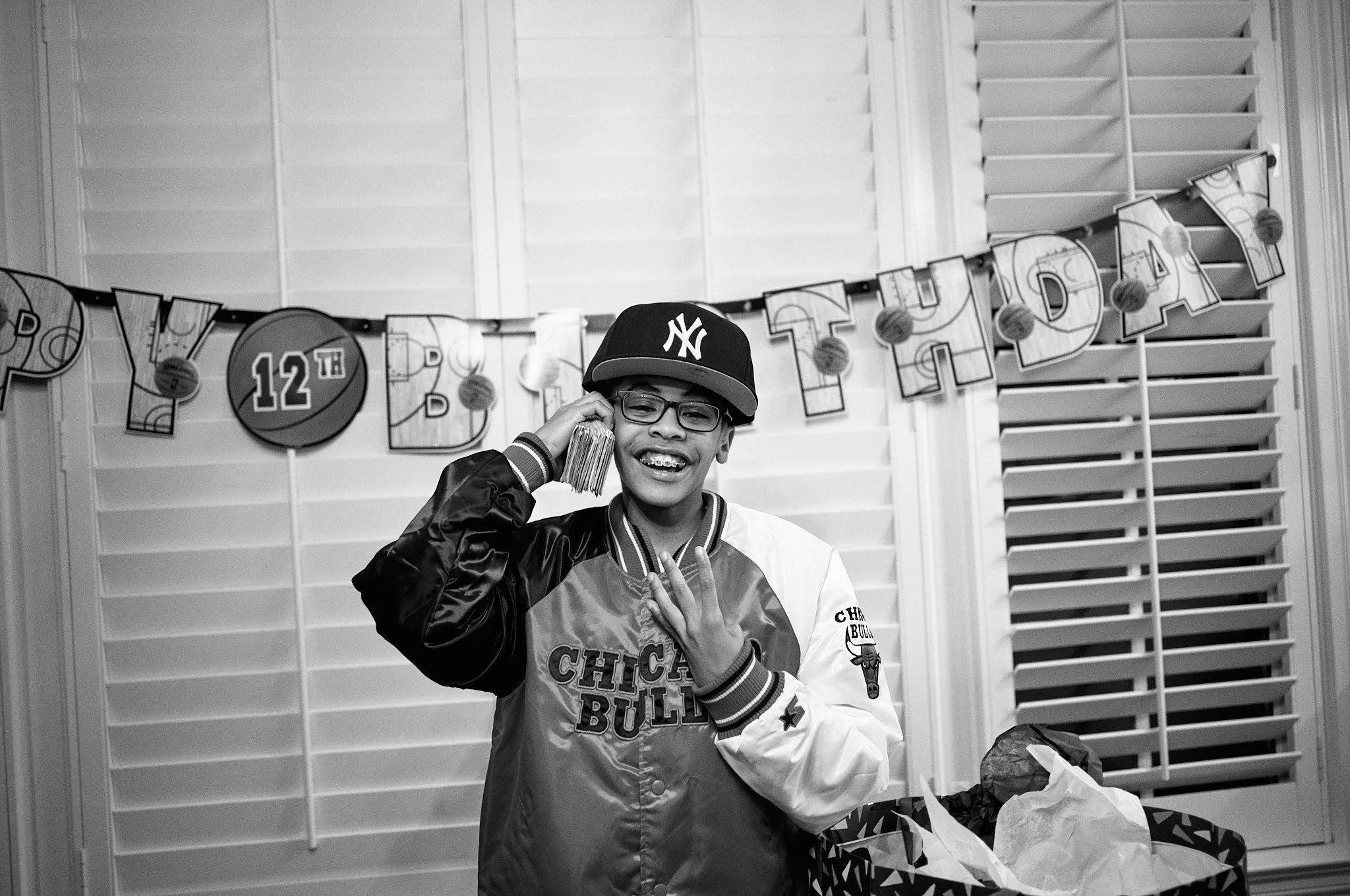 A young person celebrating their birthday at a party, wearing a Chicago Bulls jacket and a New York Yankees cap, holding a bag of money, with birthday decorations in the background.