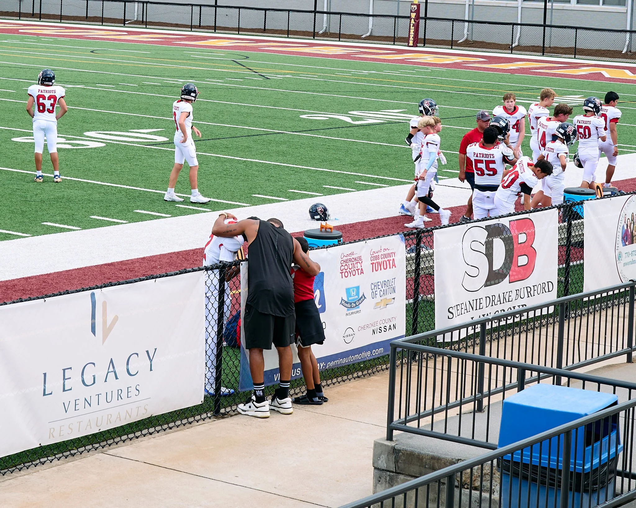 A youth football team in white and red uniforms warming up and talking on the sideline at a football field with their helmets on the ground. Two players are hugging near the fence, and coaches are nearby. Advertising banners line the fence.