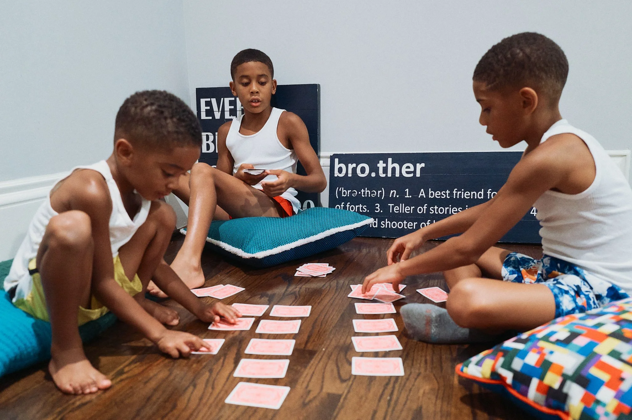 Three children playing a card game on the floor in a room with educational signs about 'bro.ther'