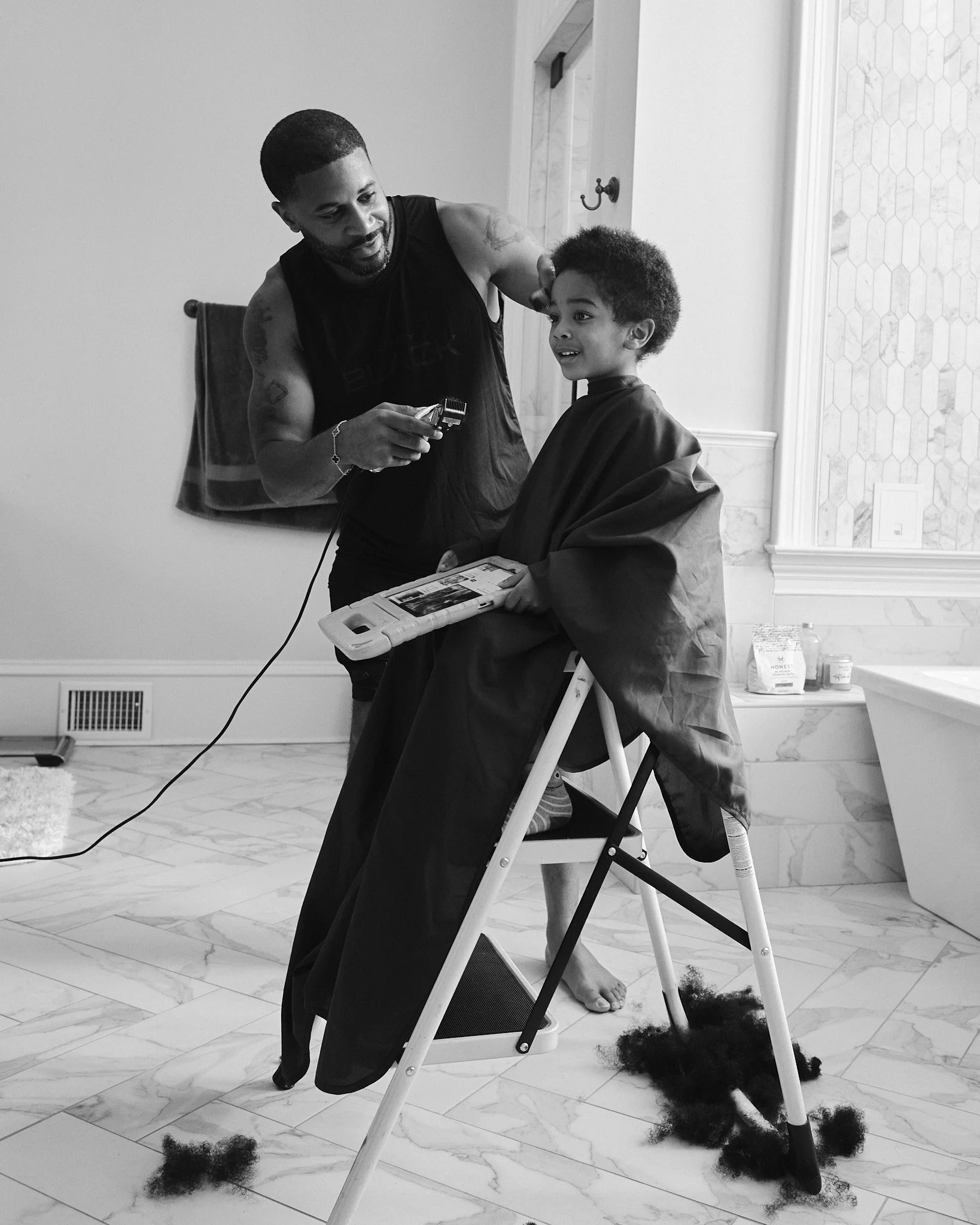 Devale Ellis giving a haircut to a young boy in a bathroom with a marble floor and a bathtub in the background, black and white photo.