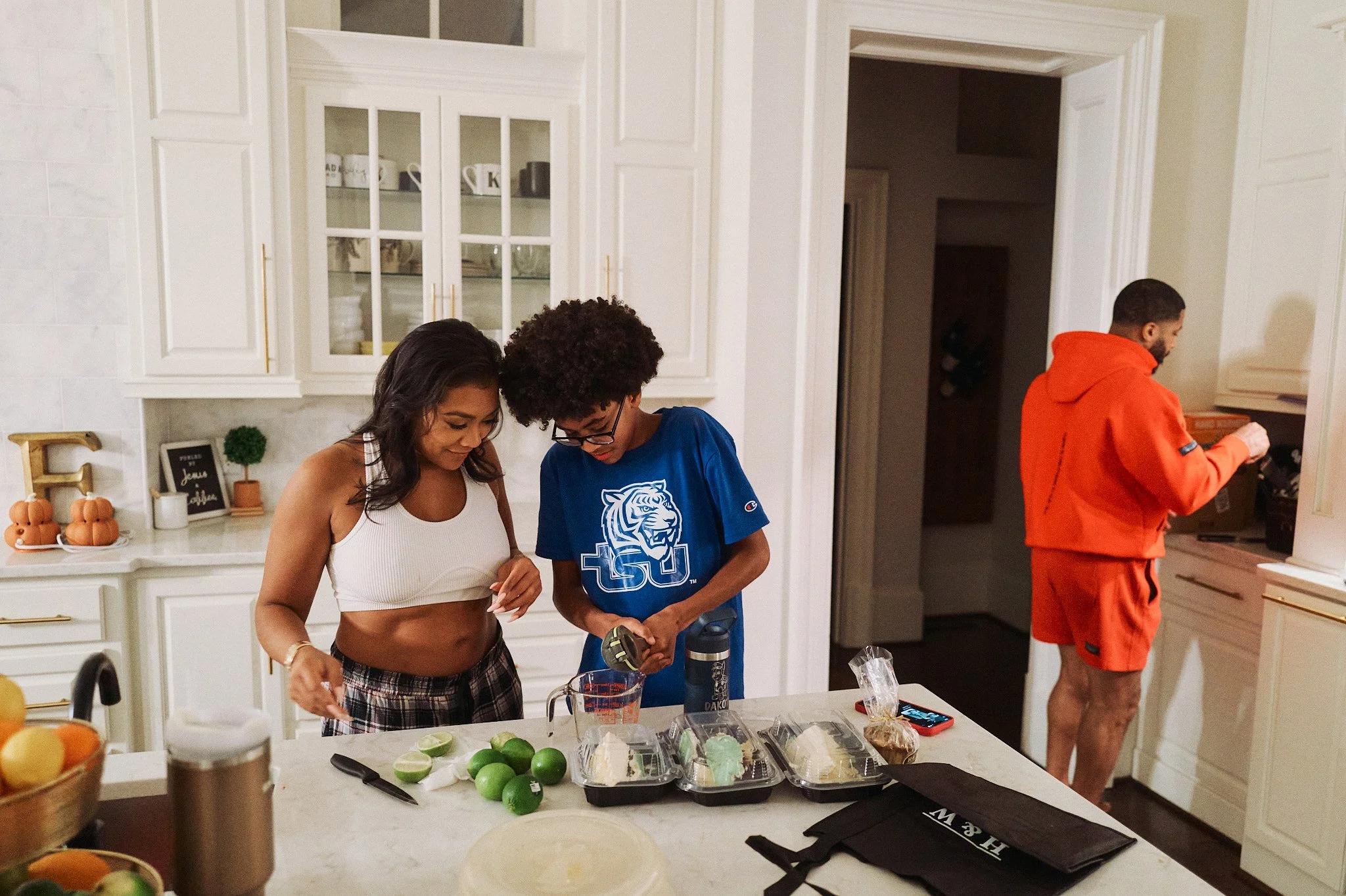 Khadeen  and Jackson preparing food in a bright kitchen, with a man in an orange outfit in the background near the counter.