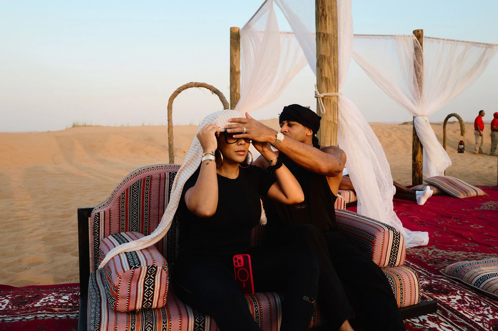 A man and woman sitting on a patterned sofa on the sand dunes of a desert, with white curtains and wooden posts around them. The man is adjusting a hat on the woman's head.