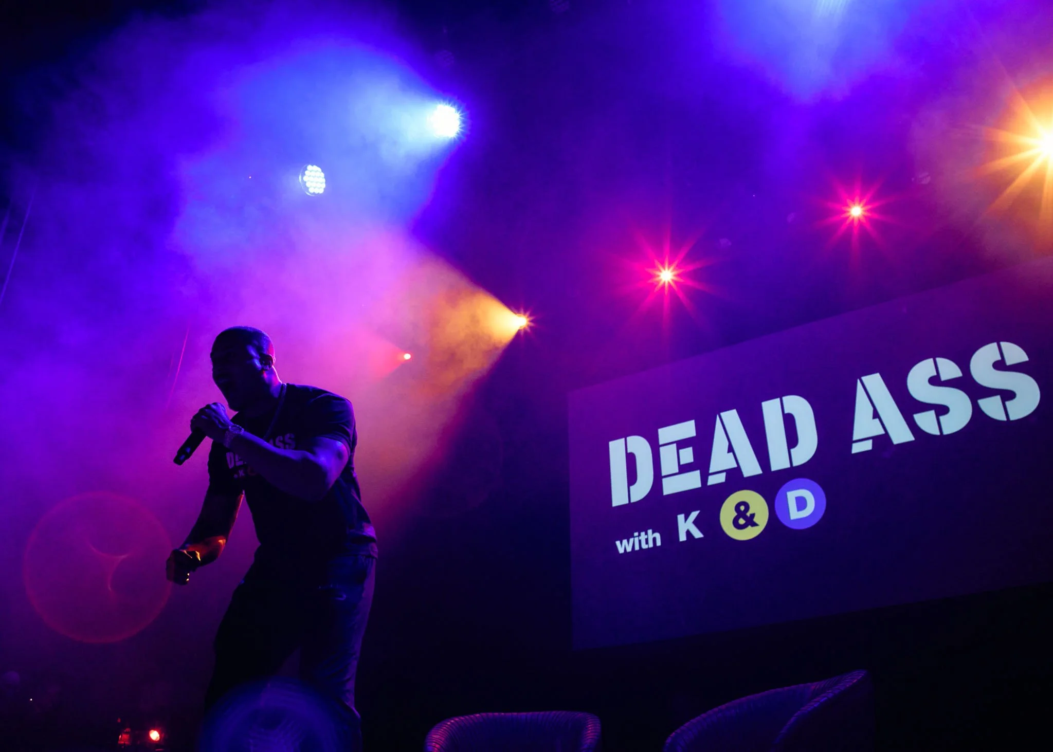 Devale holding a microphone on a stage with colorful purple, yellow, and pink lights, with a large screen displaying 'DEAD ASS with K & D' in the background.