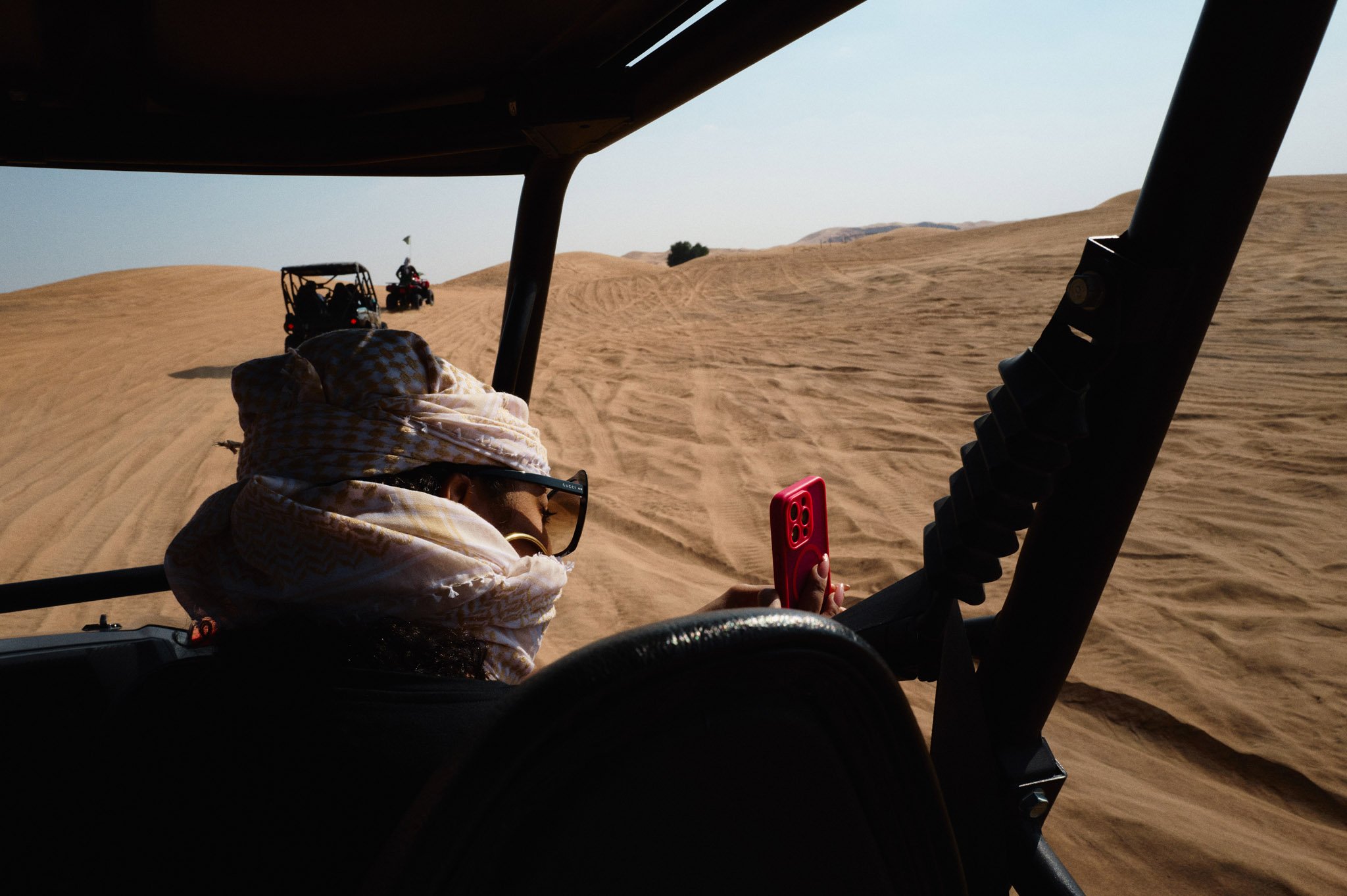 Khadeen Ellis wearing glasses and a scarf riding in a dune buggy, looking at a smartphone, in a desert with sand dunes and other dune buggies ahead.