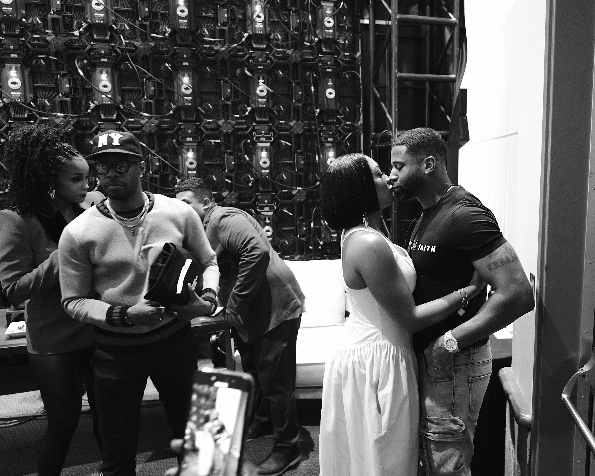 Khadeen and Devale kissing in the foreground, with several people including a woman and a man analyzing electronic equipment in the background.