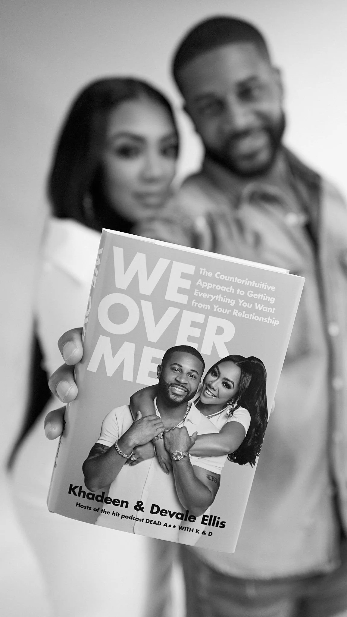 Khadeen and Devale Ellis holding a book titled "We Over Me" featuring Khadeen and Devale Ellis on the cover, with a man standing behind her, both slightly blurred in the background.
