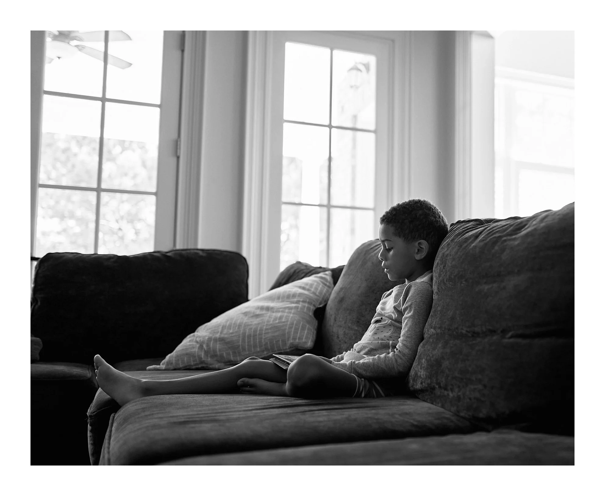 A young boy sitting on a couch, looking at a device, in a living room with large windows in the background.